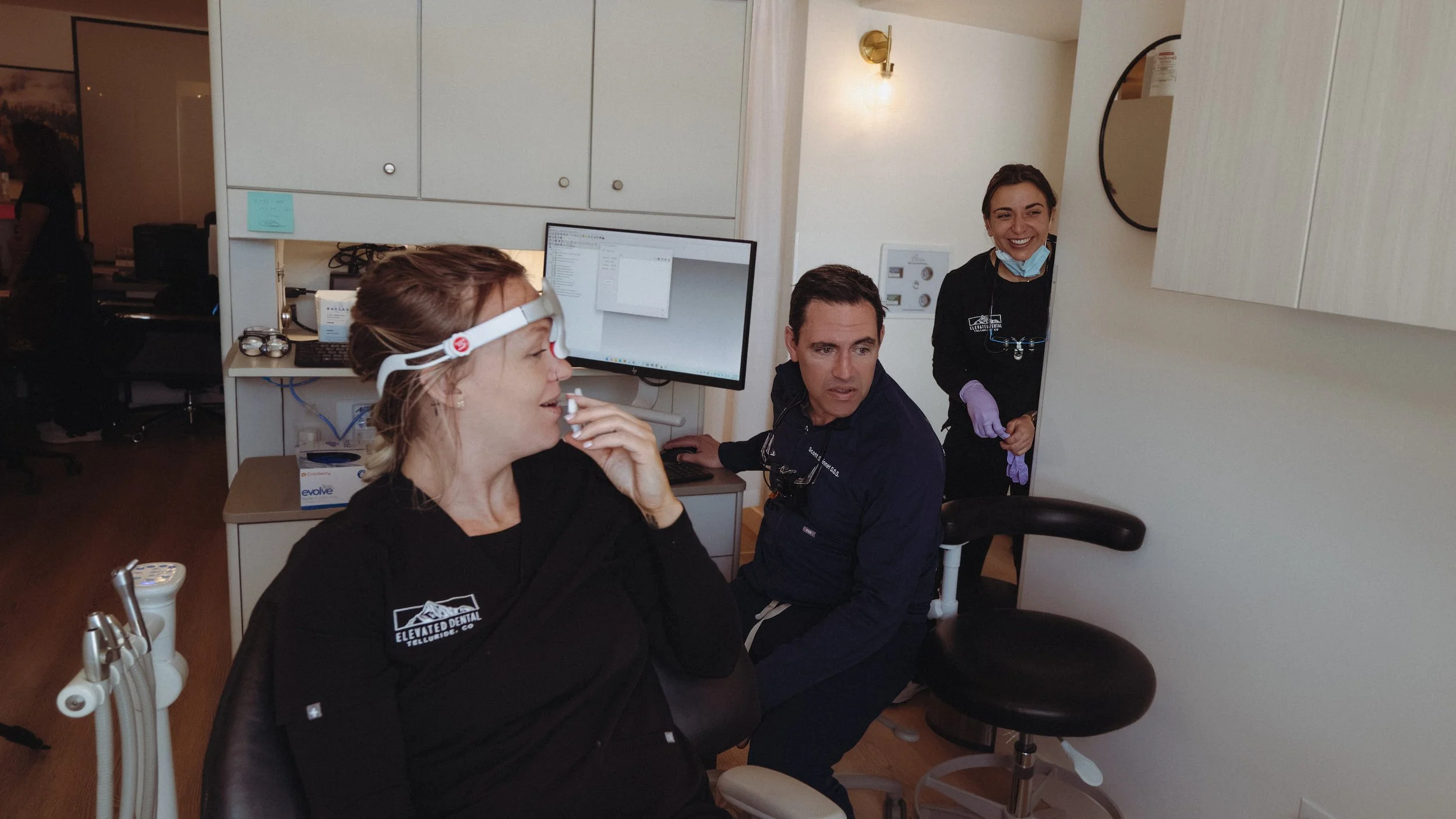 A woman in a dental chair with protective glasses, a headlamp, and a black dental gown, being examined by two dental professionals in a dental office. One professional is seated next to her, and the other is standing in the doorway, both wearing black uniforms and purple gloves. The woman appears to be comfortable and smiling.
