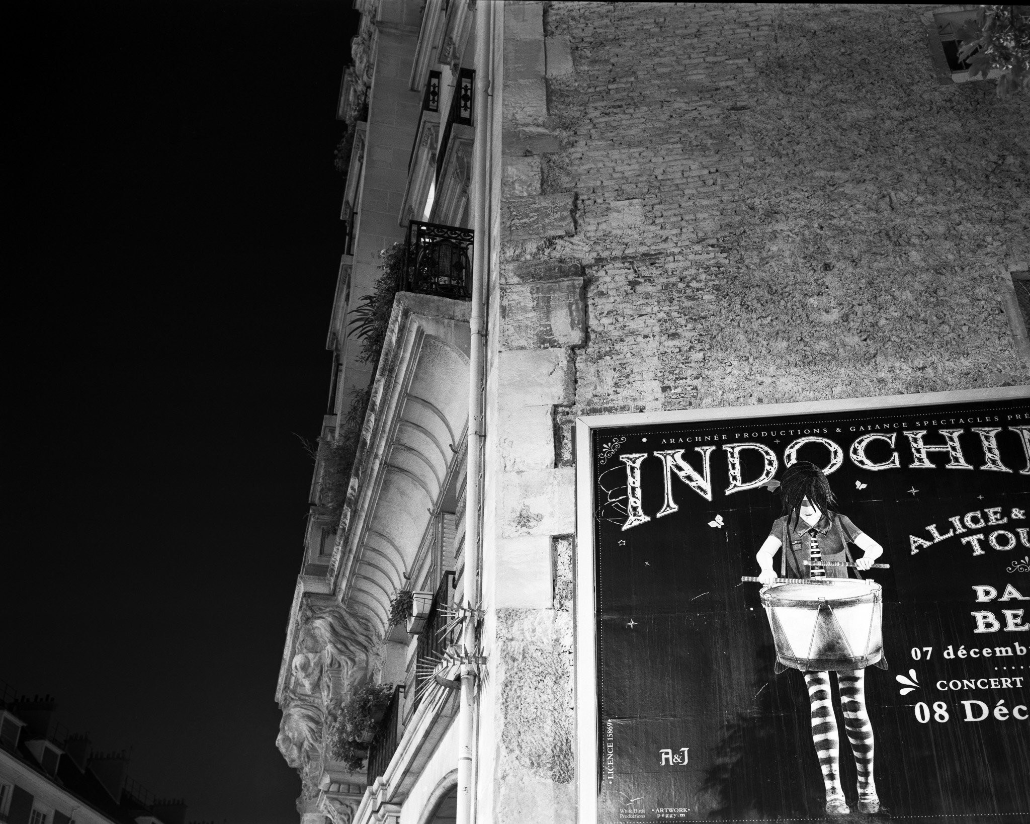 Black-and-white photograph of a nighttime street scene with a poster on a wall, Paris.