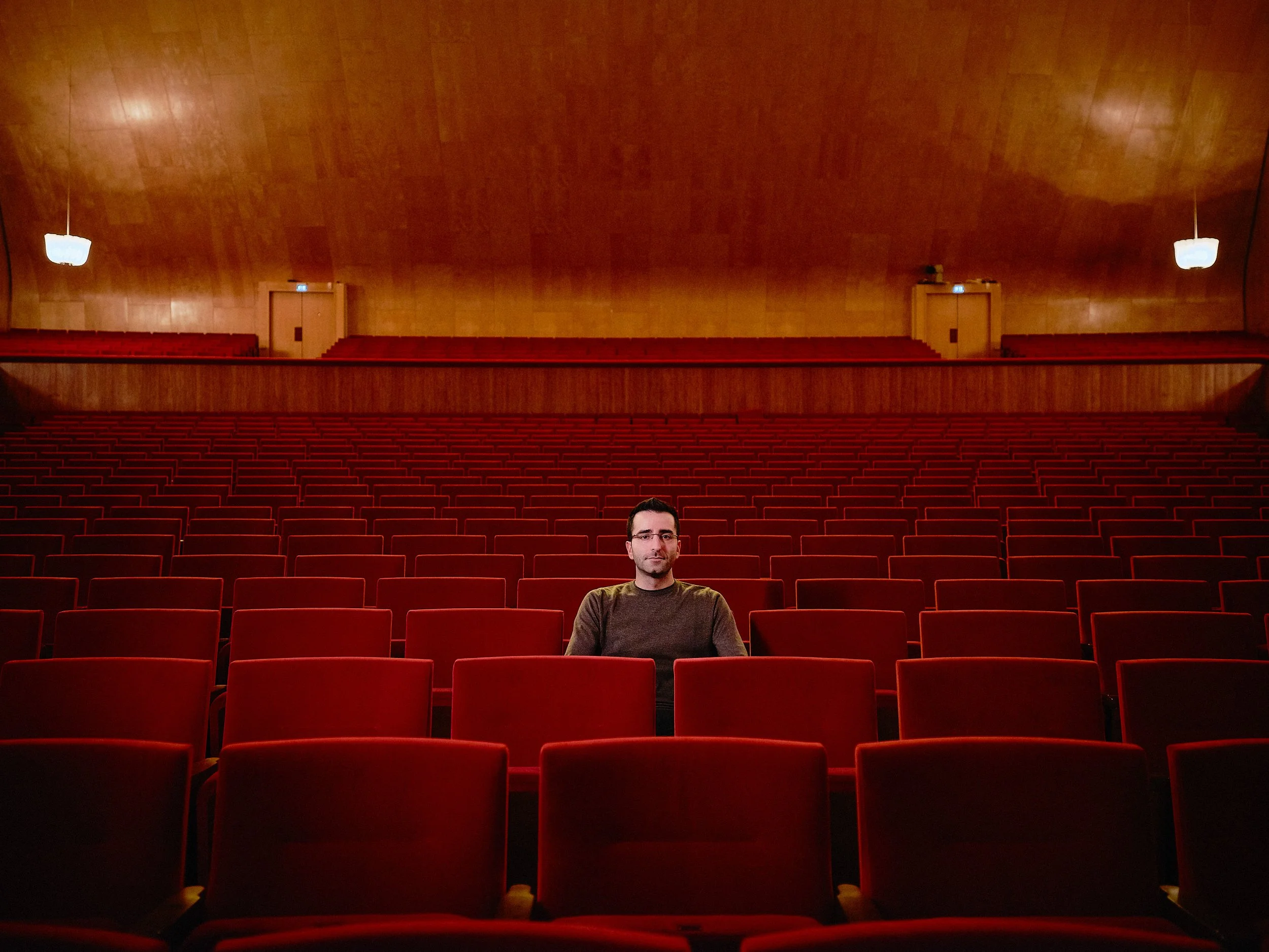 En man sitter ensam i en tom teaterrestaurang med röda stolar och trämålade väggar.