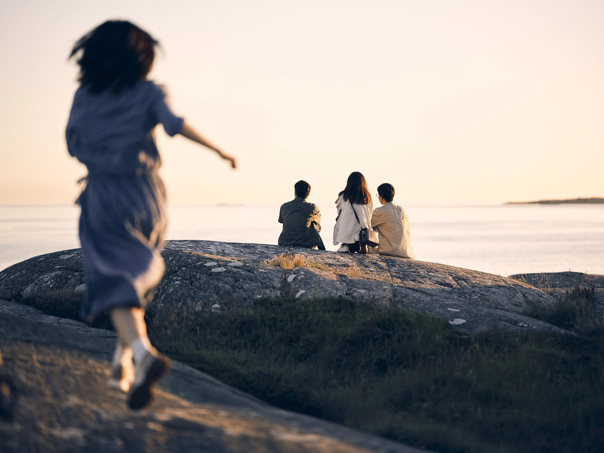 Kvinna springer mot en grupp vuxna som sitter på en sten vid vattnet under solnedgången.