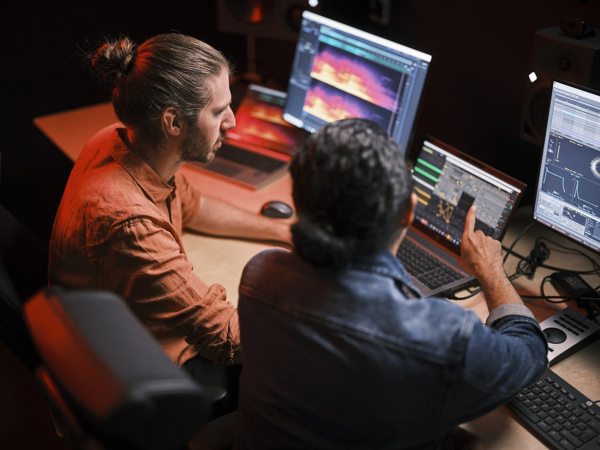Två personer arbetar med ljud- eller musikprodukter i ett studio eller kontorsmiljö med tre datorskärmar som visar ljudvågor och musikprogram.