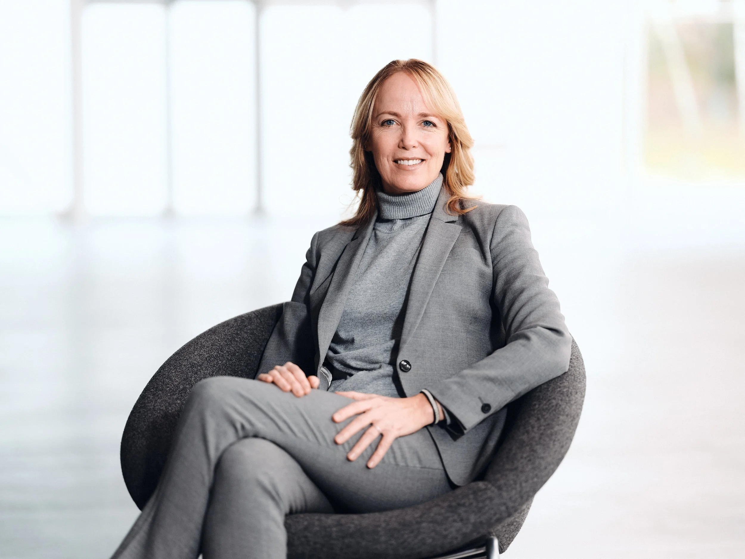 Kvinna i grå kostym sitter i en modern kontorsmiljö med stora fönster i bakgrunden.