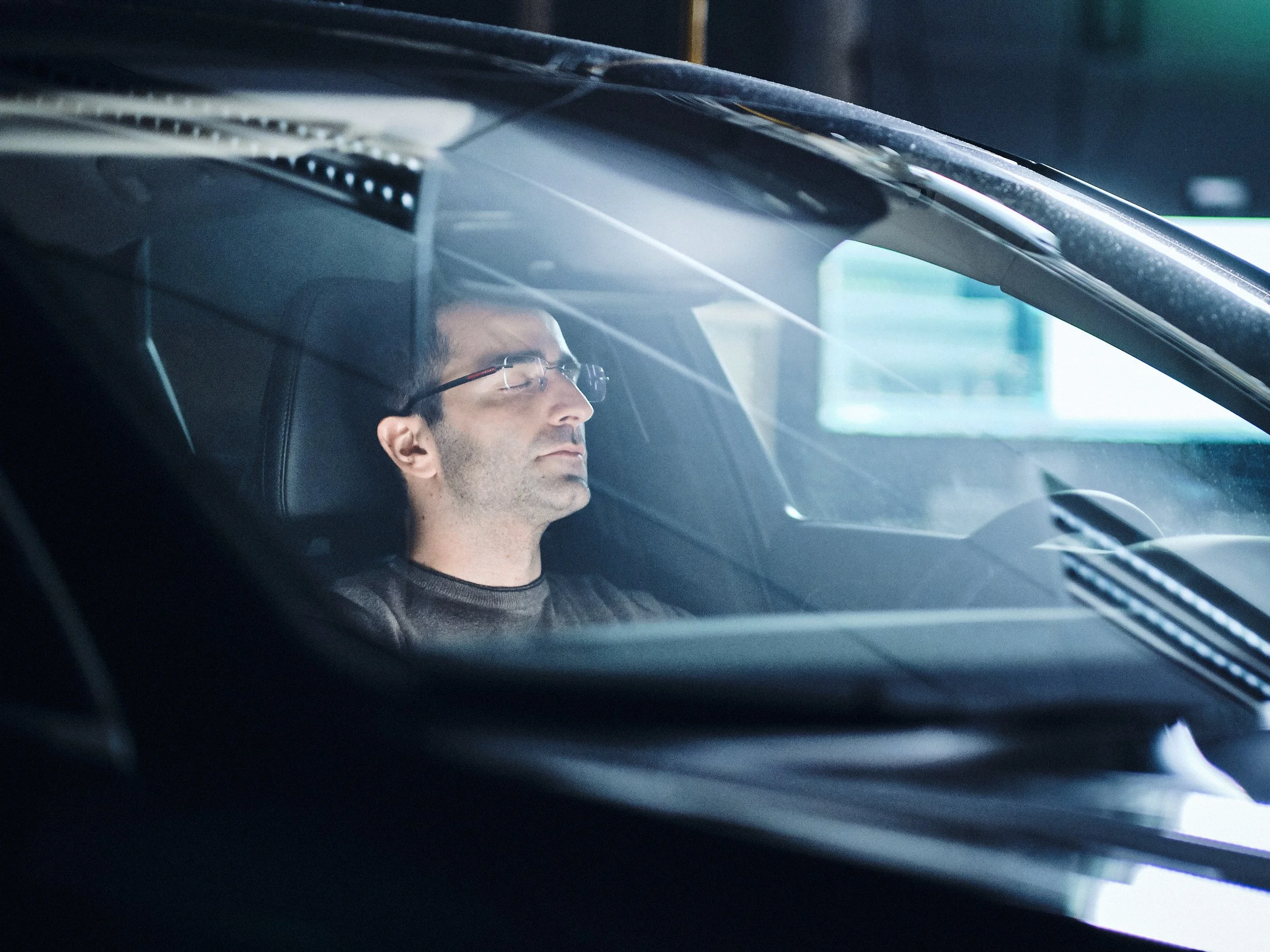 En man med glasögon som sover eller vilar sitt ansikte mot bilens säte, sett inifrån en bils vindruta.