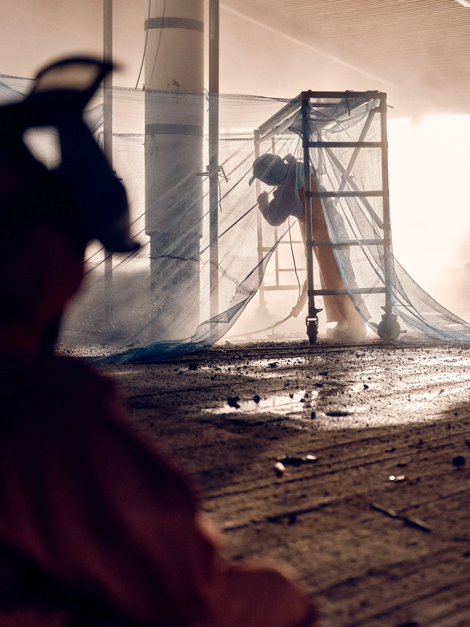 Construction worker on scaffolding working on a ceiling, with dry dirt and debris on the ground, and sunlight streaming in from the side.