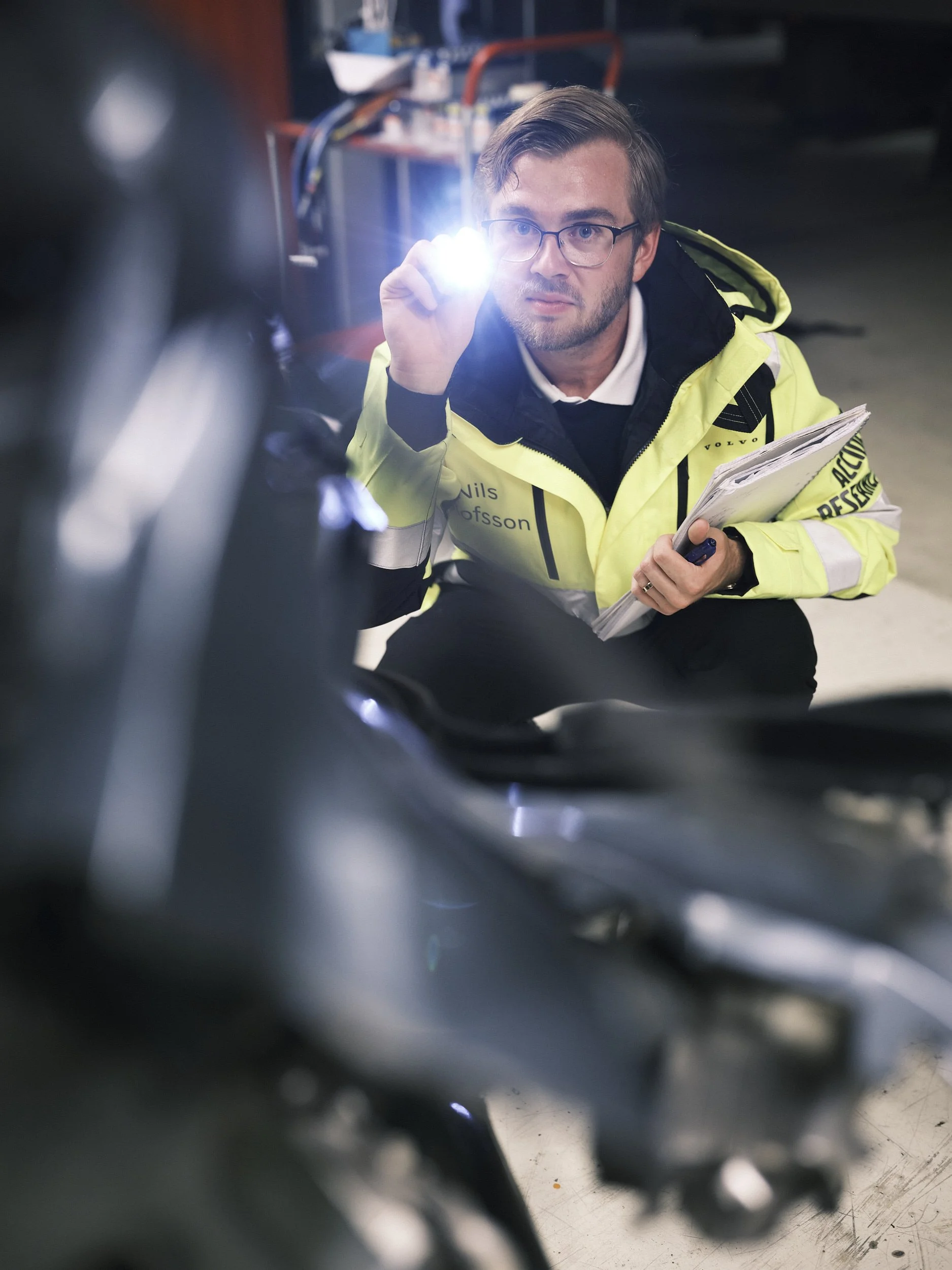 En man i en gul jacka med texten 'Nils' och 'Volvo' som håller en penna och en bunt papper, tittar ner på en maskin eller motor i ett verkstadsmiljö, medan han riktar en ficklampa mot den.