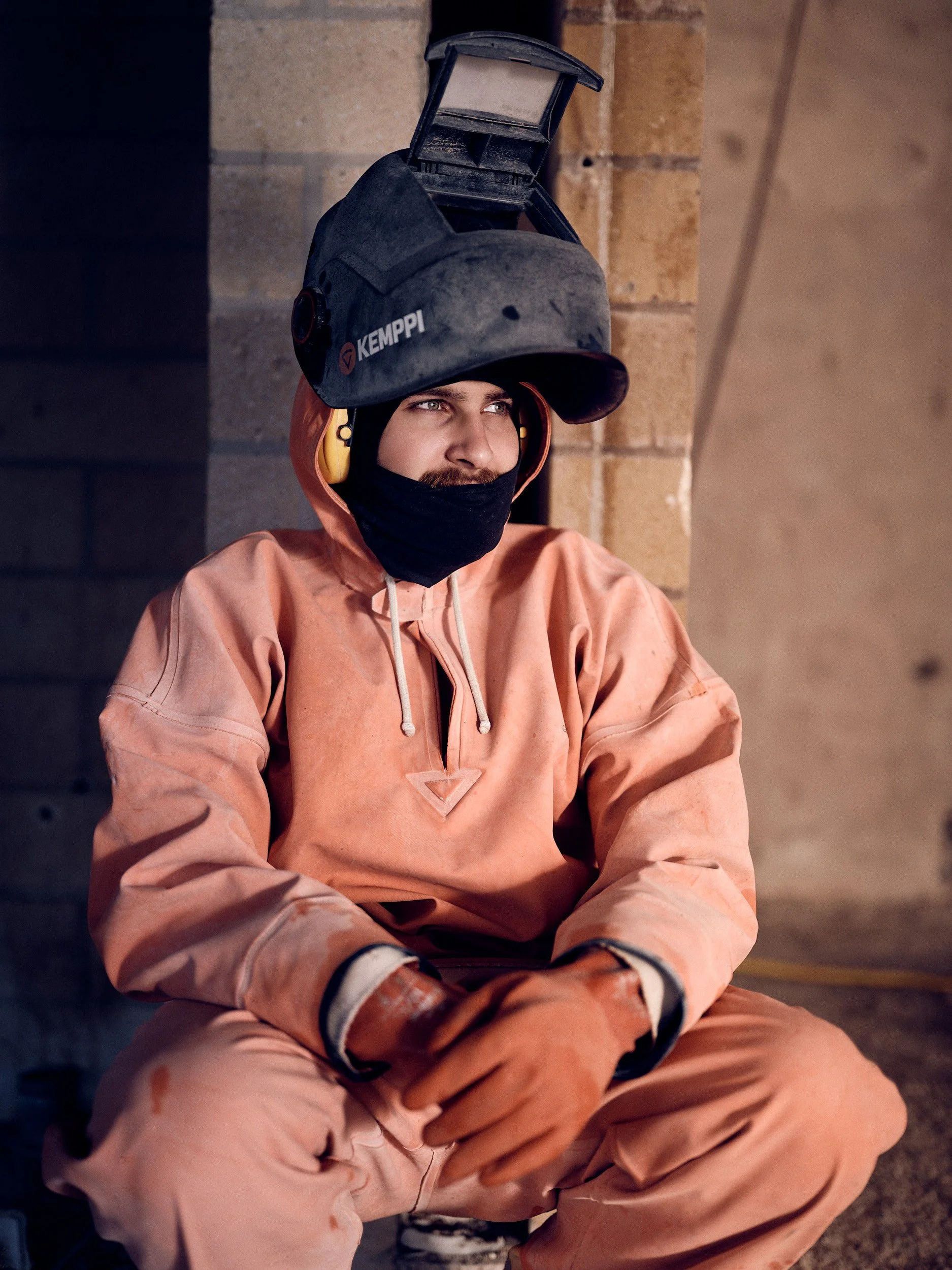 A construction worker wearing orange protective gear, gloves, and a black face mask, sitting against a wall with a welding helmet on his head.