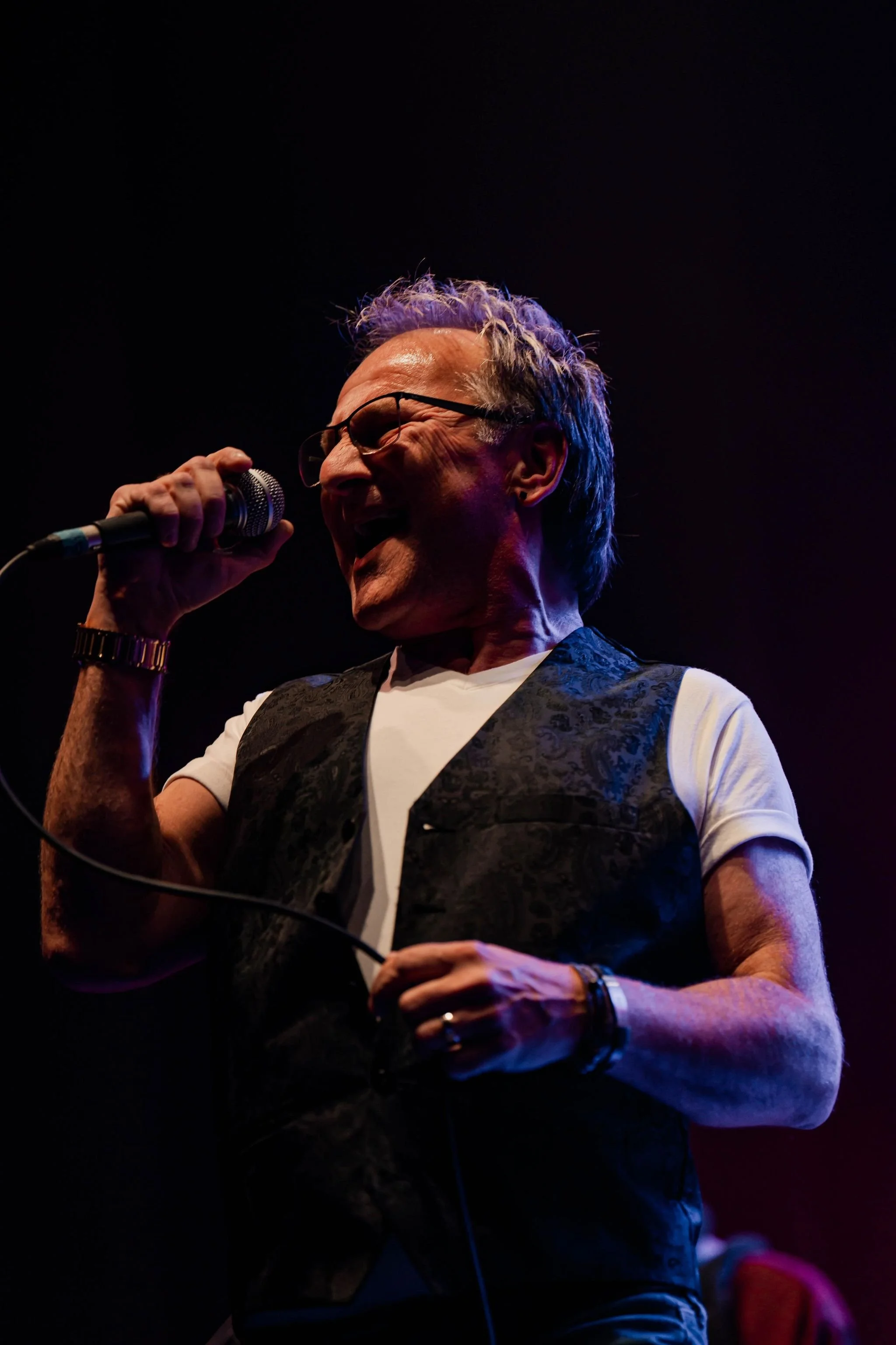 A man with glasses and gray hair singing into a microphone on stage, dressed in a white t-shirt and a dark vest.