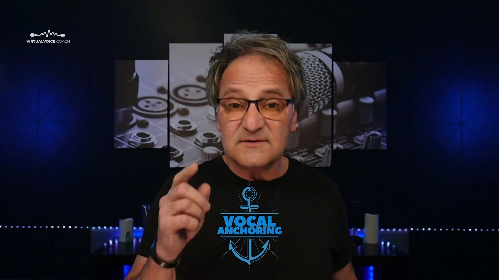 A man with glasses and gray hair pointing his finger forward in a vocal coaching studio with a dark background, blue LED lights, and a backdrop featuring a microphone and audio equipment. The words "VOCAL ANCHORING" are printed on his black shirt with a nautical anchor logo.