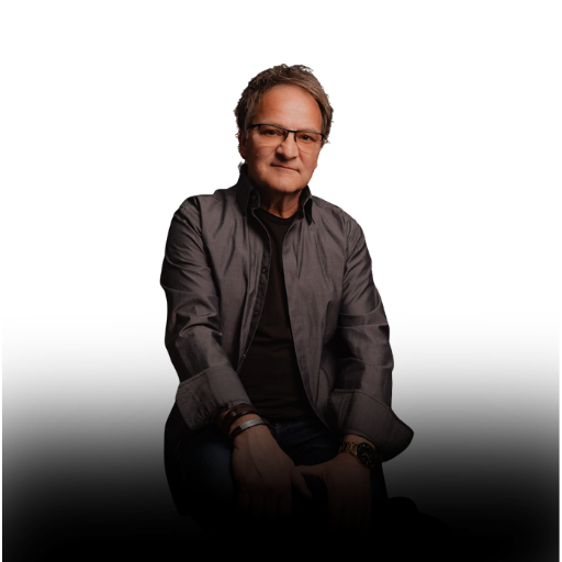 A middle-aged man wearing glasses, a dark shirt, and a gray jacket, sitting with a neutral expression against a black background.