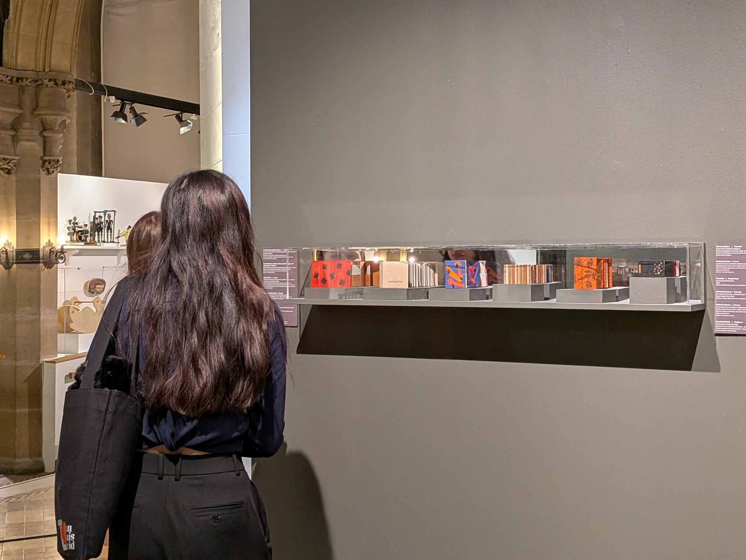 Vue sur une vitrine de l'exposition Small Wonders avec deux personnes de dos la contemplant, permettant de donner une échelle aux livres et comprendre la scénographie