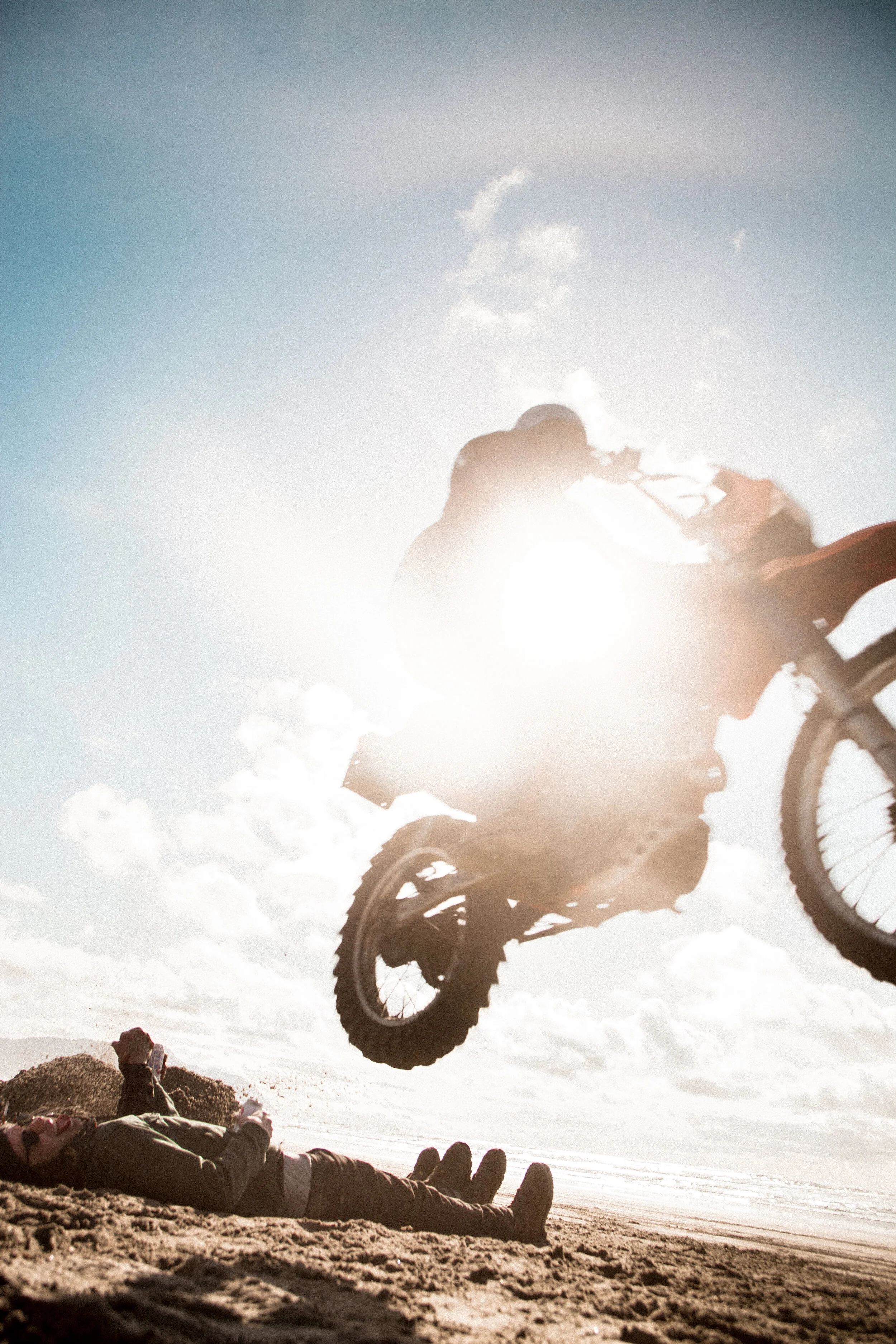 A dirt bike jumps over a person lying on a sandy beach, the sun shining brightly and creating a dramatic silhouette against the sky. Photographed by Will Corwin.