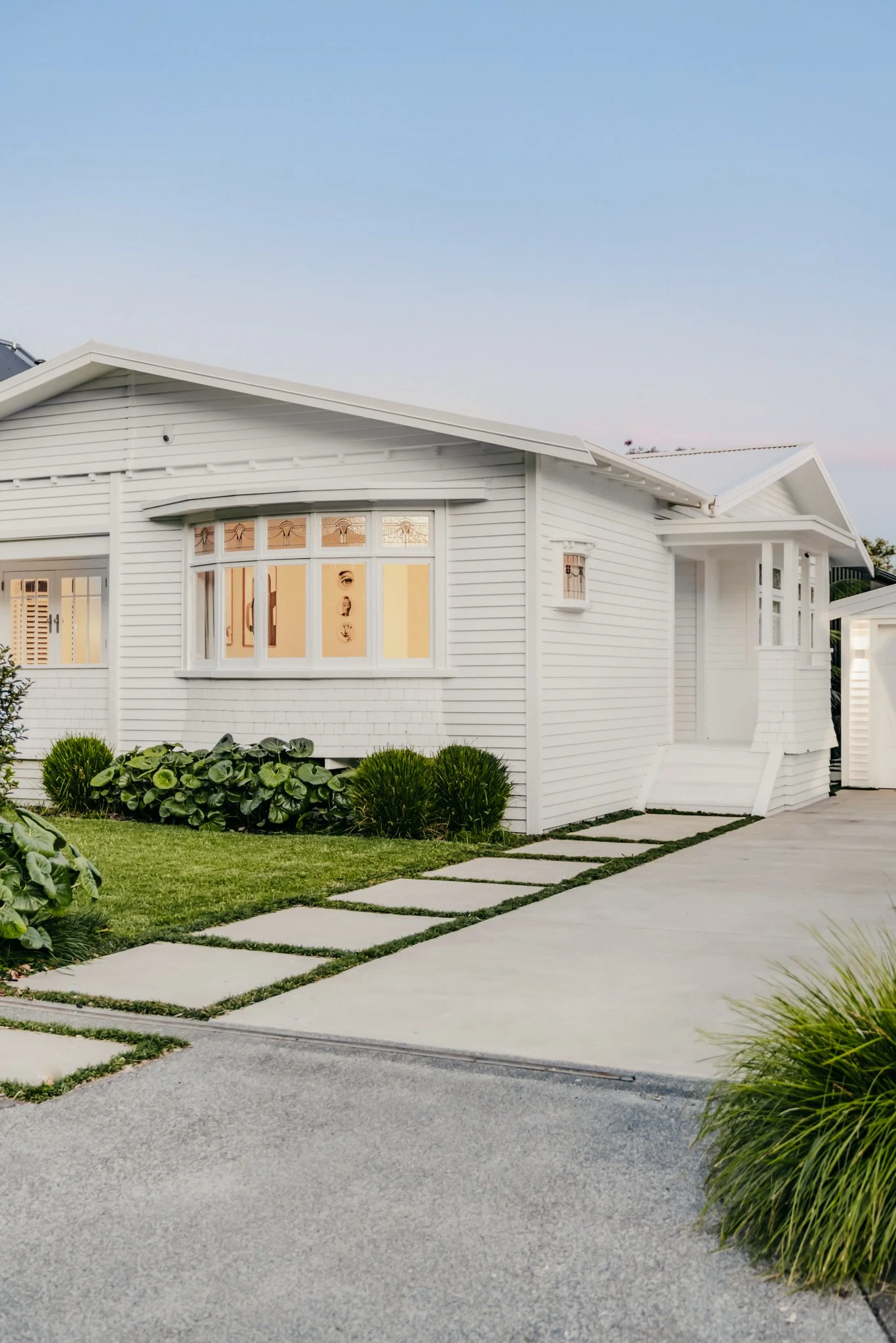 Street frontage of the Point Chevalier Transitional Bungalow by Macfie Architecture at dusk, with restored box bay window, leadlight glazing and white weatherboard cladding