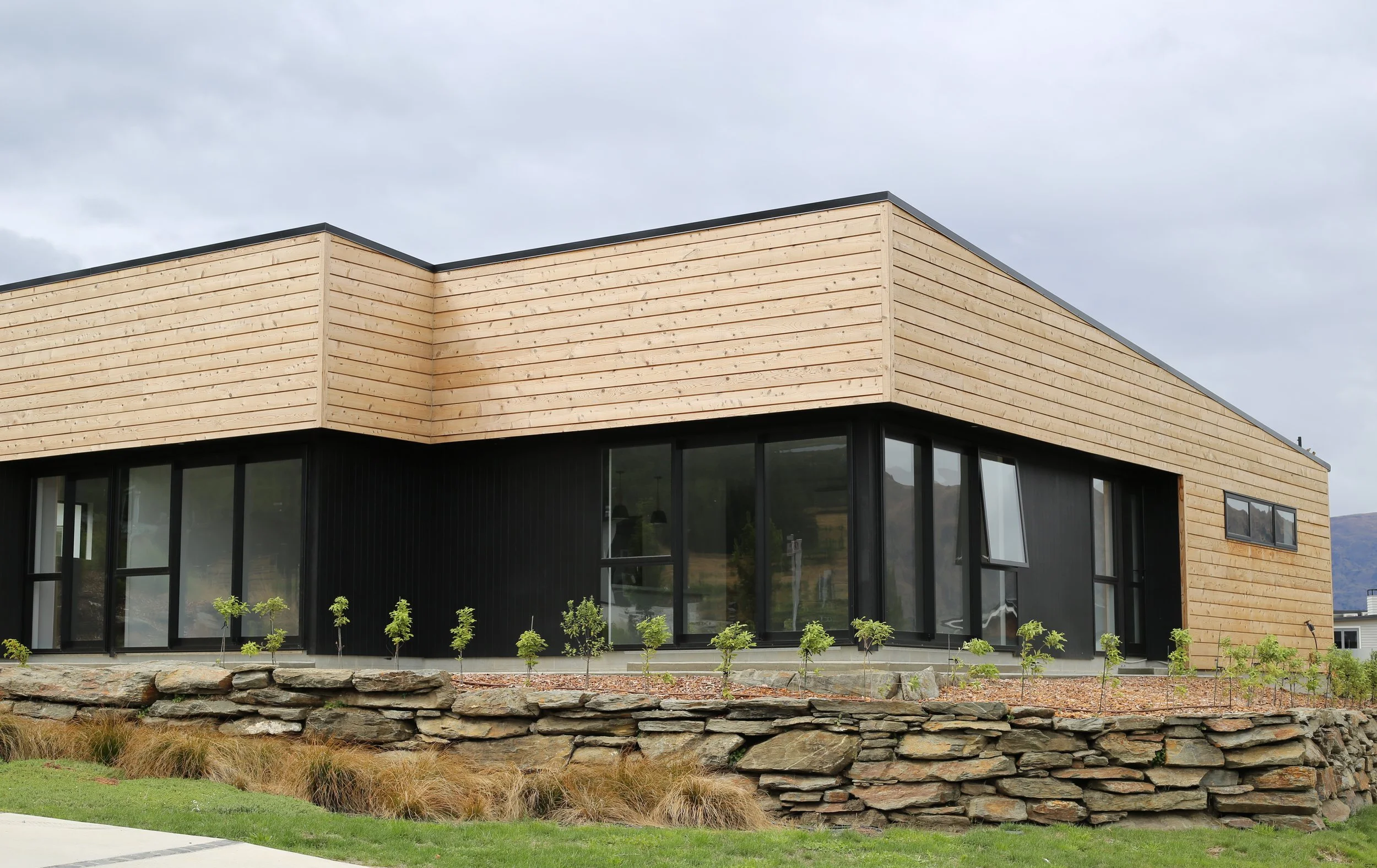 Wanaka new-build exterior with blonde cedar weatherboard, black vertical cladding and black aluminium joinery above a schist dry-stone retaining wall