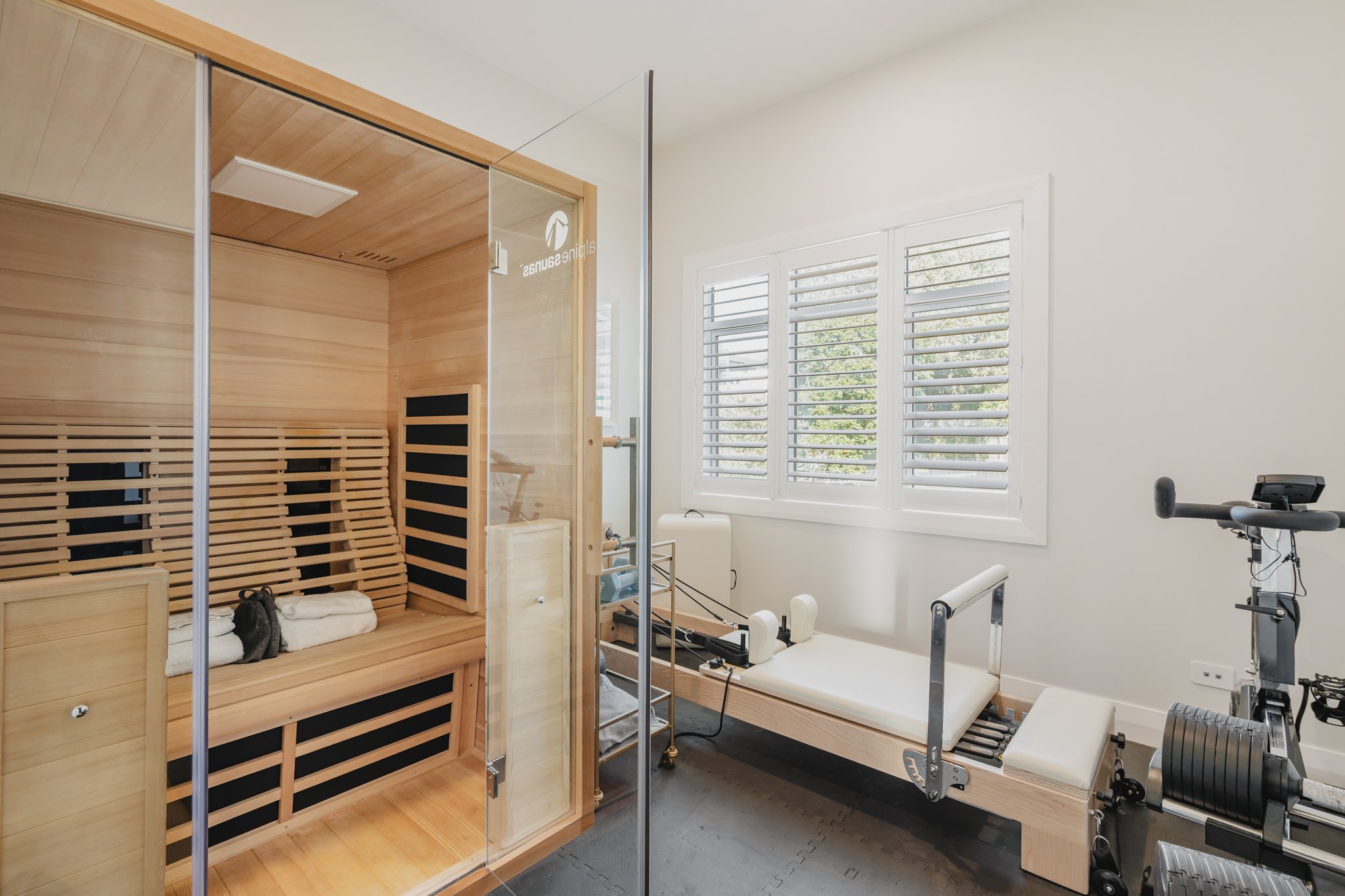 Home gym in a Westmere new build bungalow, featuring a cedar infrared sauna with glass door, pilates reformer, exercise bike, plantation shutters and rubber flooring, located in the upstairs master suite level.