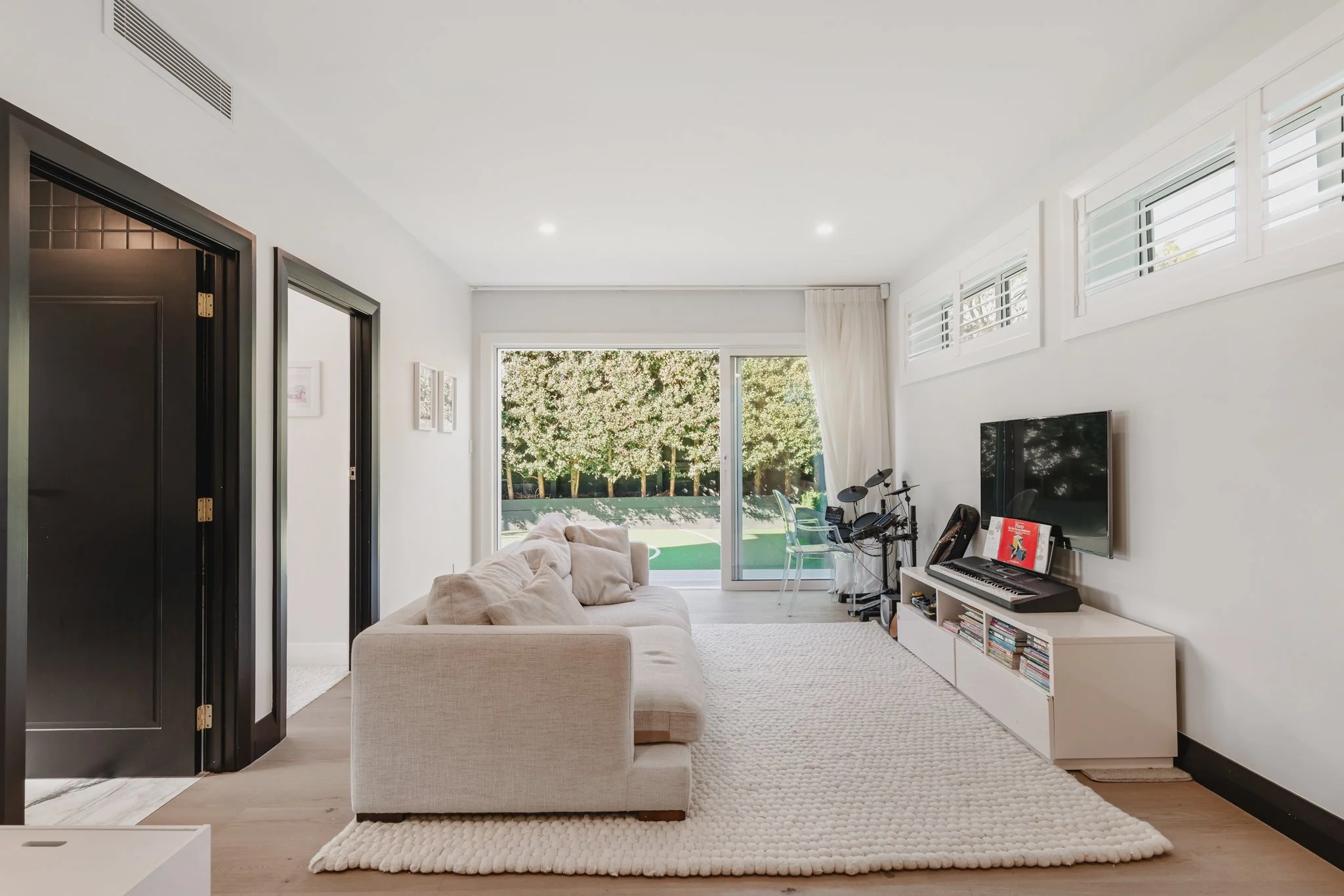 Children's lounge in a Westmere new build bungalow, featuring a neutral sectional sofa, drum kit, keyboard and television, plantation shutters, sliding doors opening directly to a backyard sports court, and black painted heritage-style door joinery.