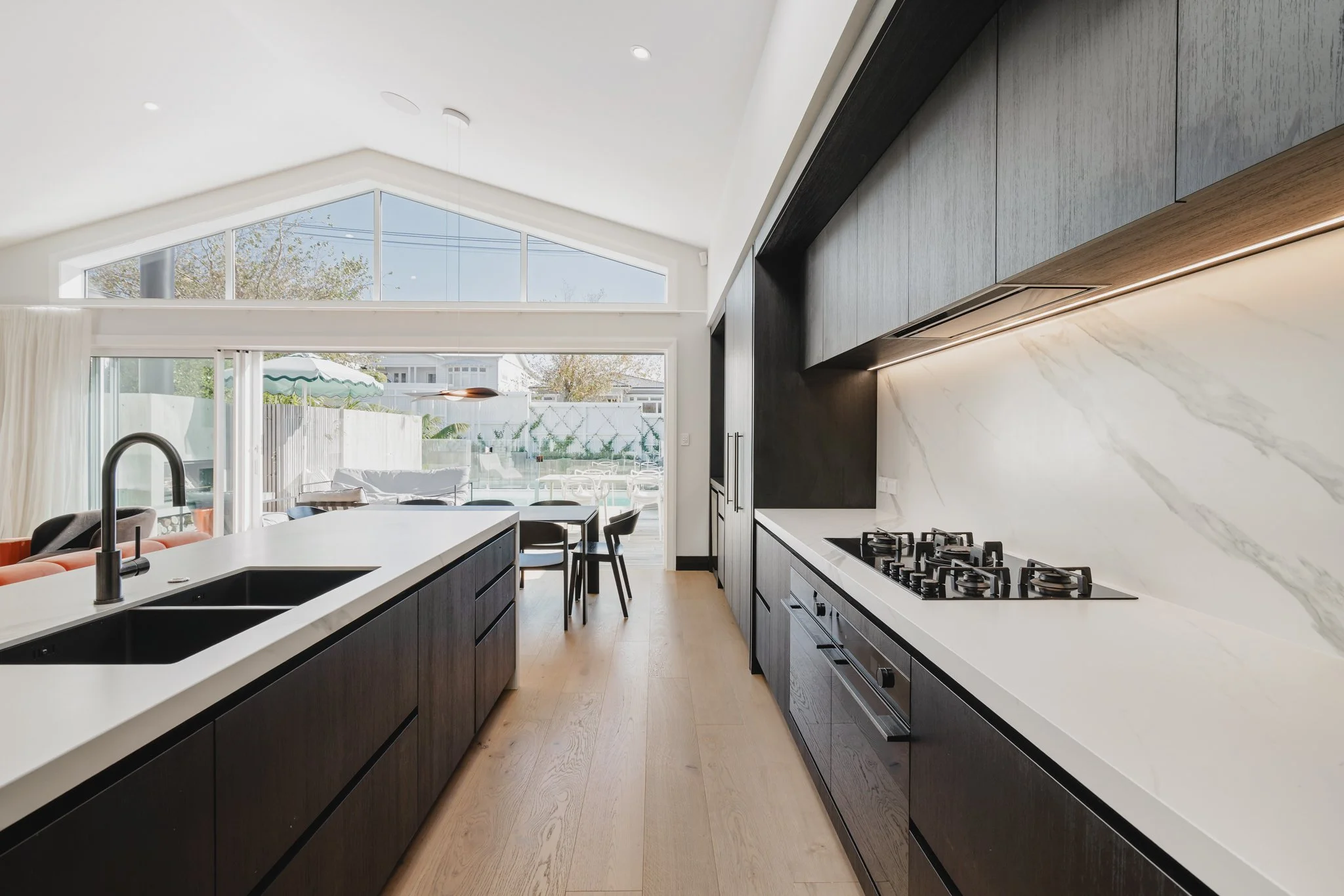 Galley kitchen with dark oak joinery, marble benchtop, gas hob and matte black tapware in a new build Auckland bungalow, with north-facing outlook to pool and entertaining terrace, Westmere