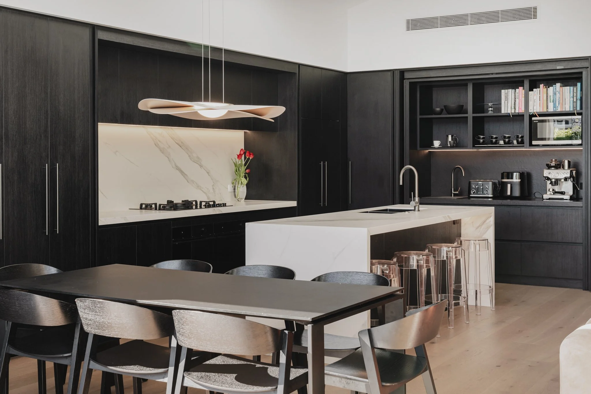 Kitchen and dining in Westmere new build bungalow, featuring floor-to-ceiling dark oak cabinetry, marble stone counter and splash backs, and sculptural pendant light black dining table and chairs and hidden butlers pantry with open shelving.