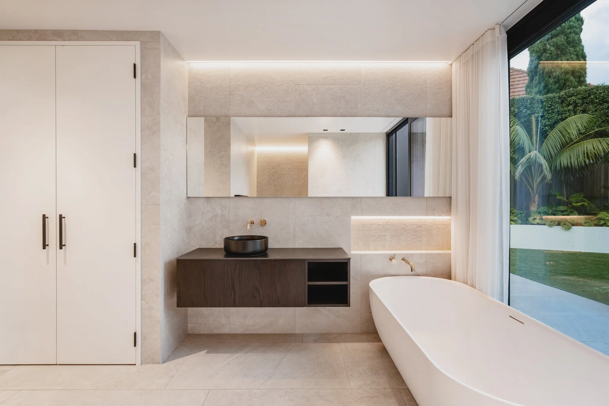 Bespoke new-build property with spa like bathroom, soft ambient LED lighting frames the natural stone bathroom with a neutral palette and dark oak cabinetry and polished brass accents.