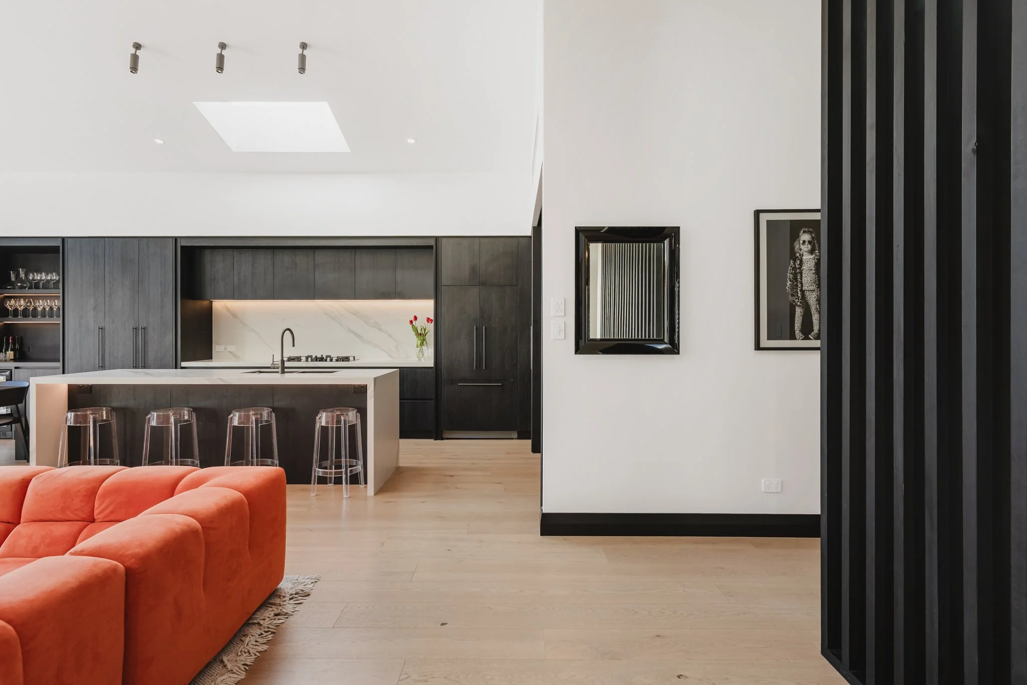 Open plan living and kitchen in a Westmere new build bungalow, showing dark oak floor-to-ceiling cabinetry, marble island bench, skylight, oak timber floors, and a bold black vertical batten stair screen with framed artwork beyond.