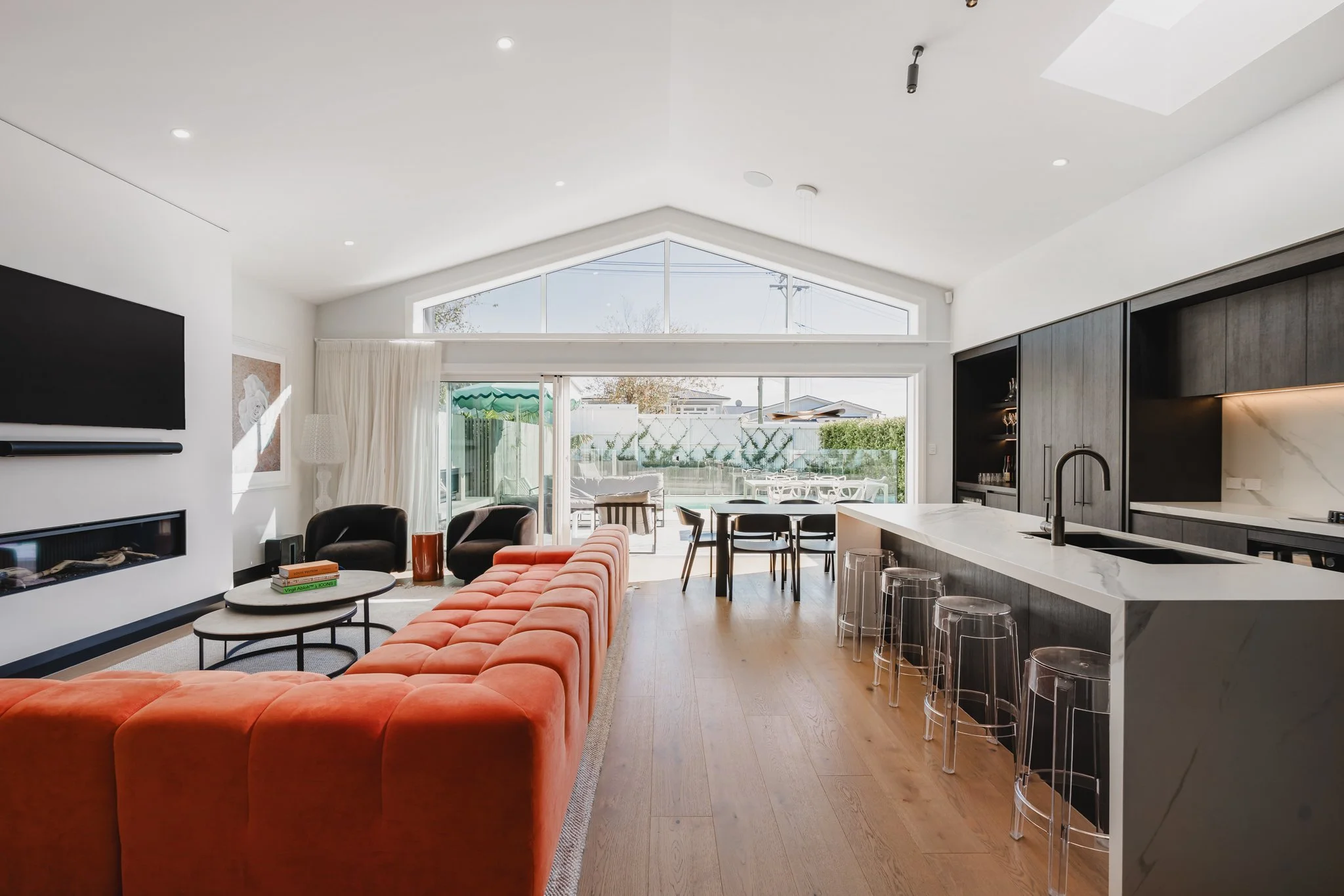 North-facing open plan living, dining and kitchen in a new build Westmere bungalow, featuring a vaulted gabled glass wall, dark oak joinery, marble island bench, and bold orange sectional sofa opening to a pool and entertaining terrace.