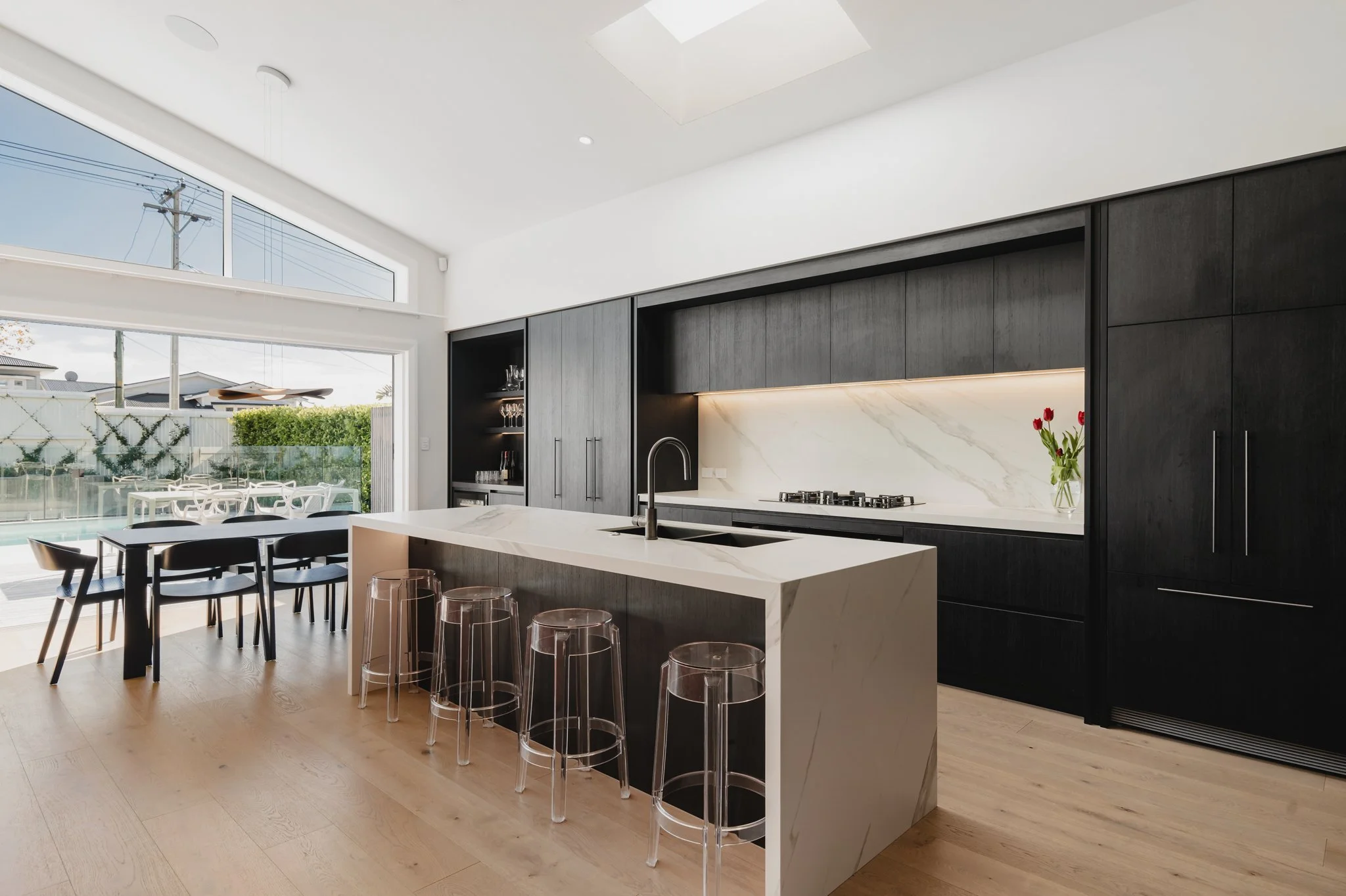 North-facing kitchen in a Westmere new build bungalow, featuring a large marble stone island bench with acrylic bar stools, floor-to-ceiling dark oak cabinetry, marble splashback with LED underlighting, gas hob, skylight, and open sliding doors.