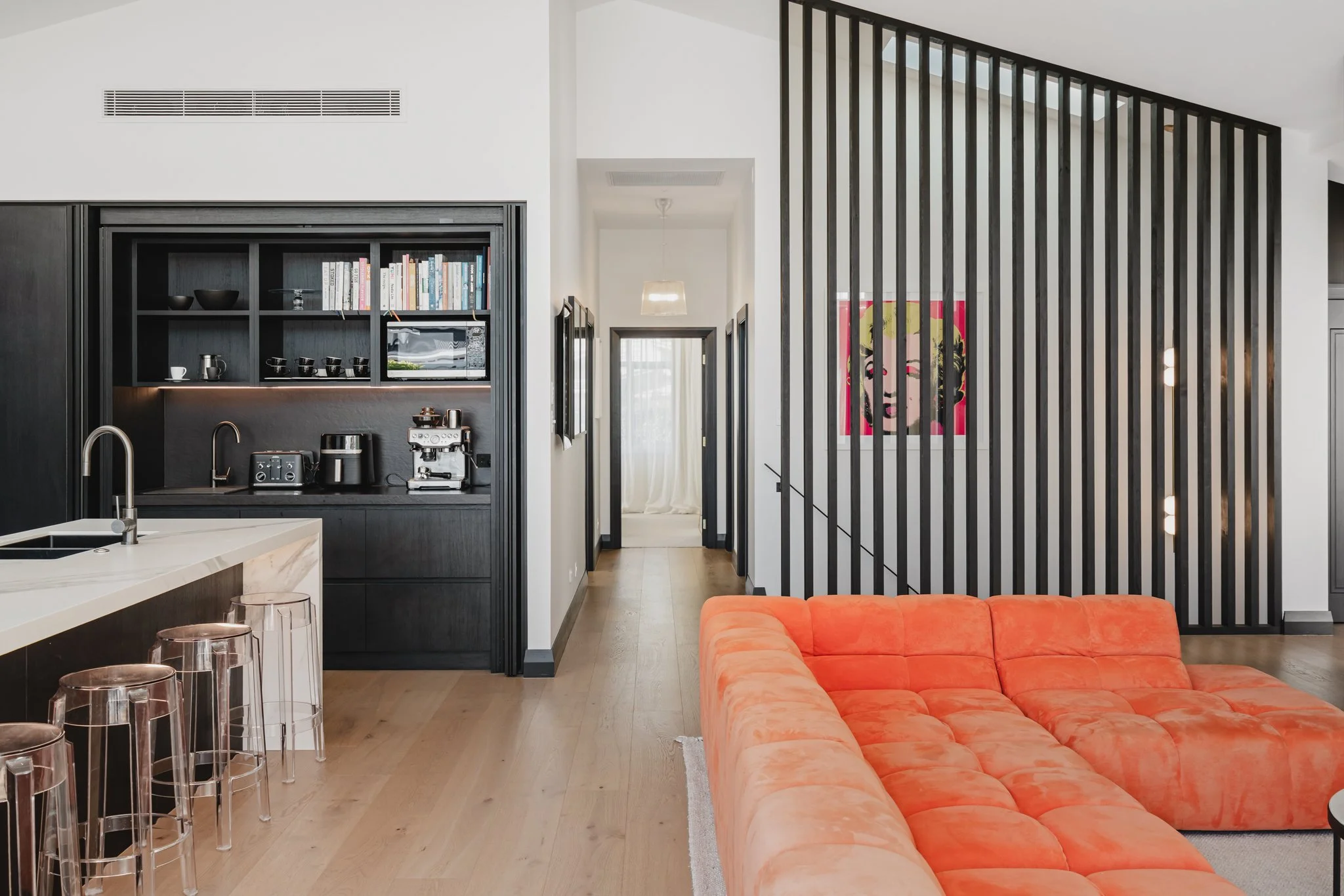 Living area and butler's pantry in a Westmere new build bungalow, showing a bold orange sectional sofa, black vertical batten stair screen with pop art, dark oak open shelving with coffee station, marble island bench.