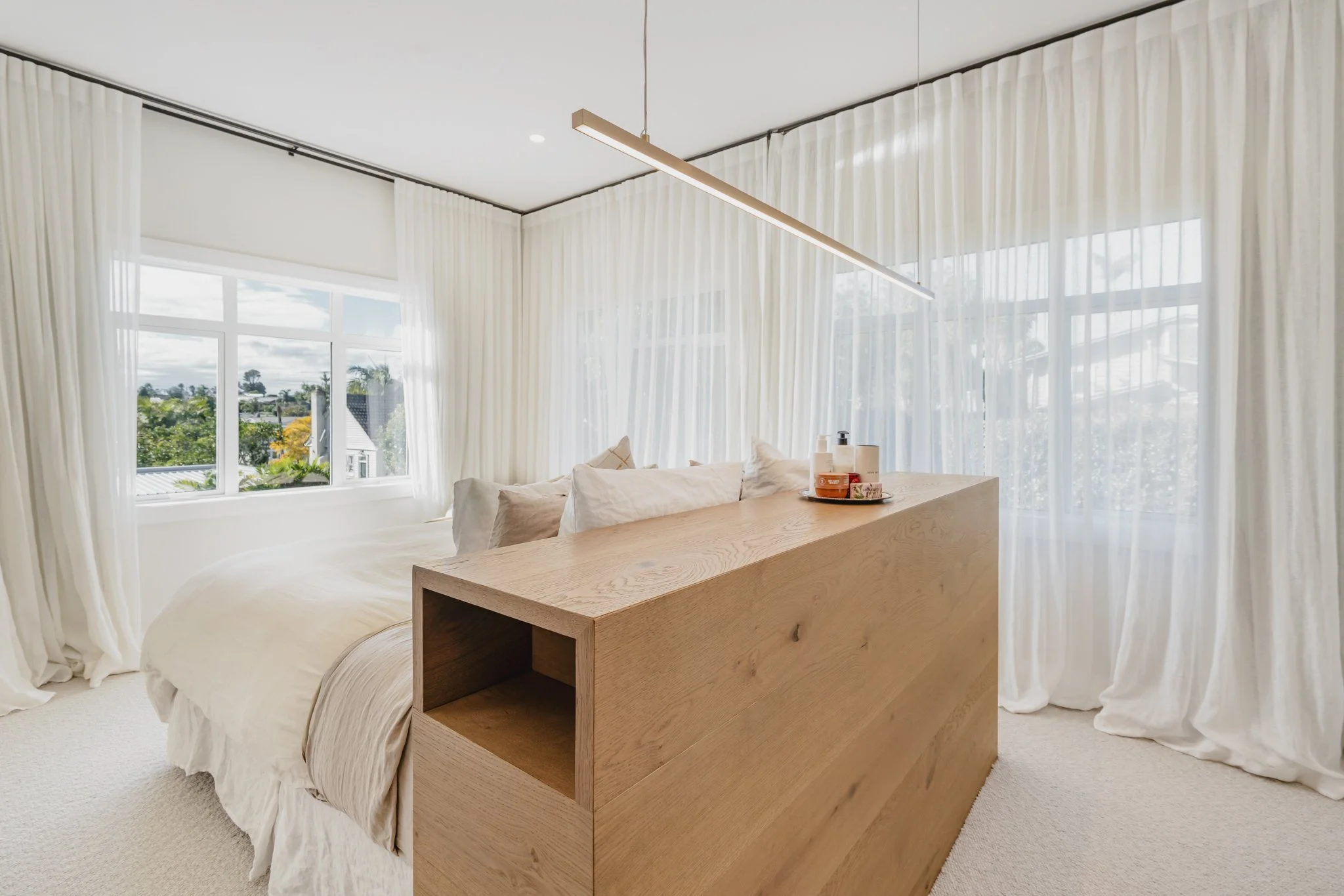 Master bedroom with bespoke oak bed surround, floor-to-ceiling sheer curtains and linear pendant light in a new build Auckland bungalow, Westmere.