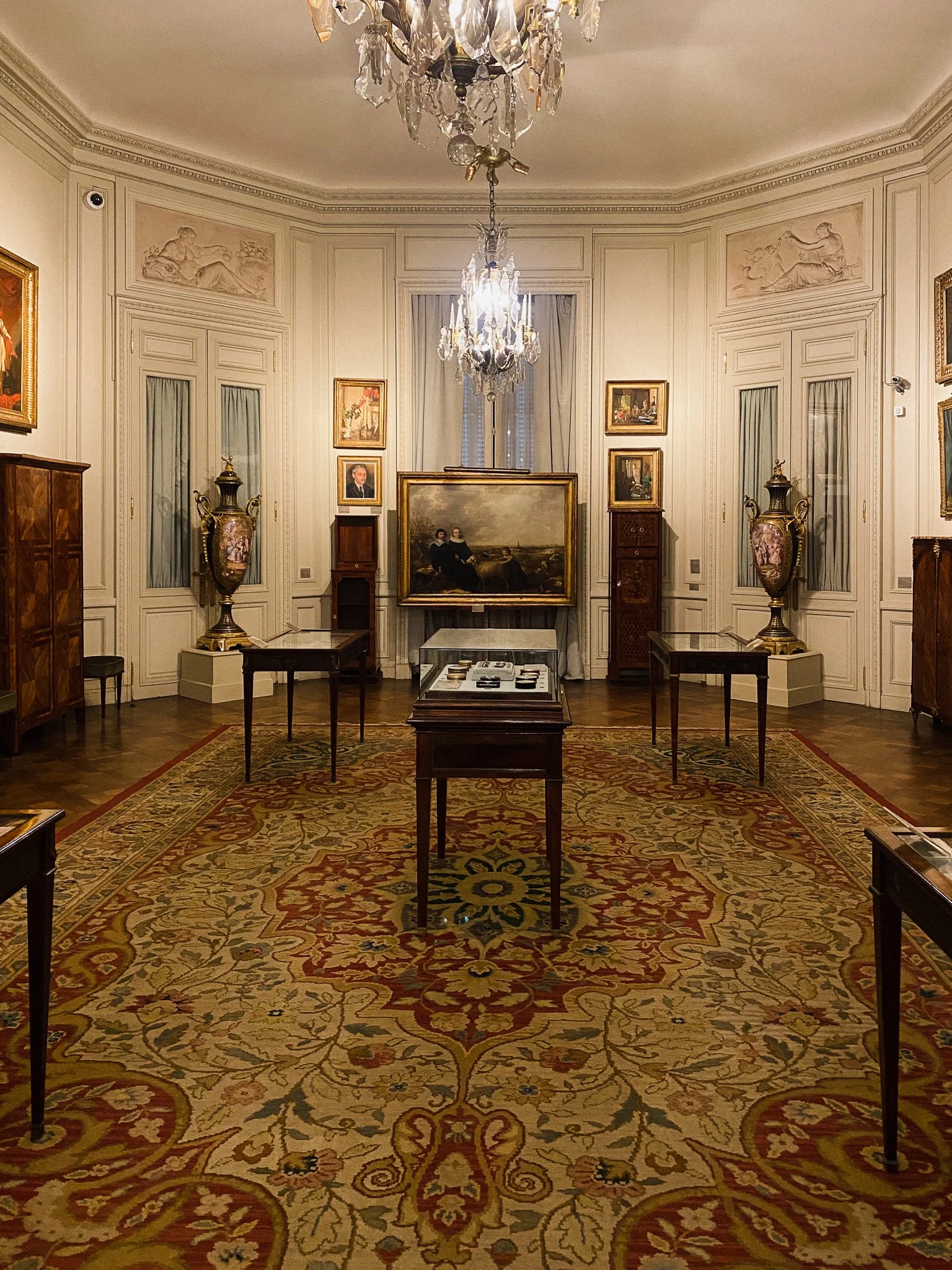 A room at the Museo Nacional de Arte Decorativo in Buenos Aires. The room is decorated with a large persian carpet, ornate millwork, paintings, ceramics, and furnishings.
