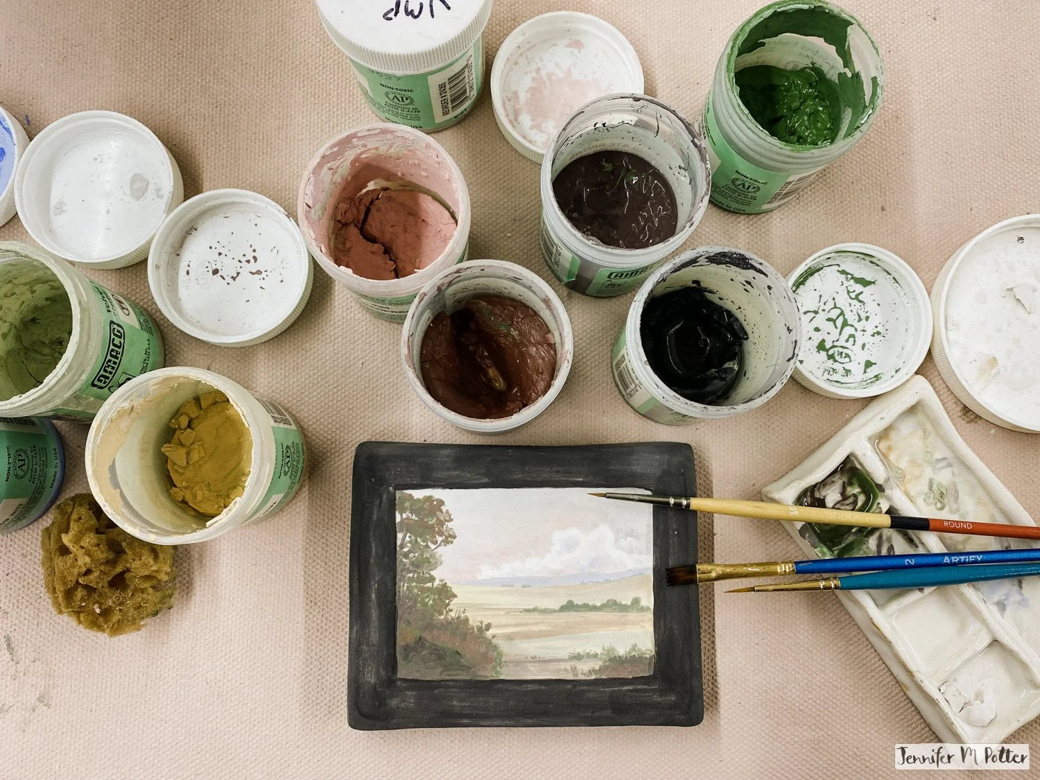 A small unfired landscape painting in underglaze on a bisque framed canvas. It is surrounded by small jars of underglaze, paintbrushes and a small hand-build watercolor palette.