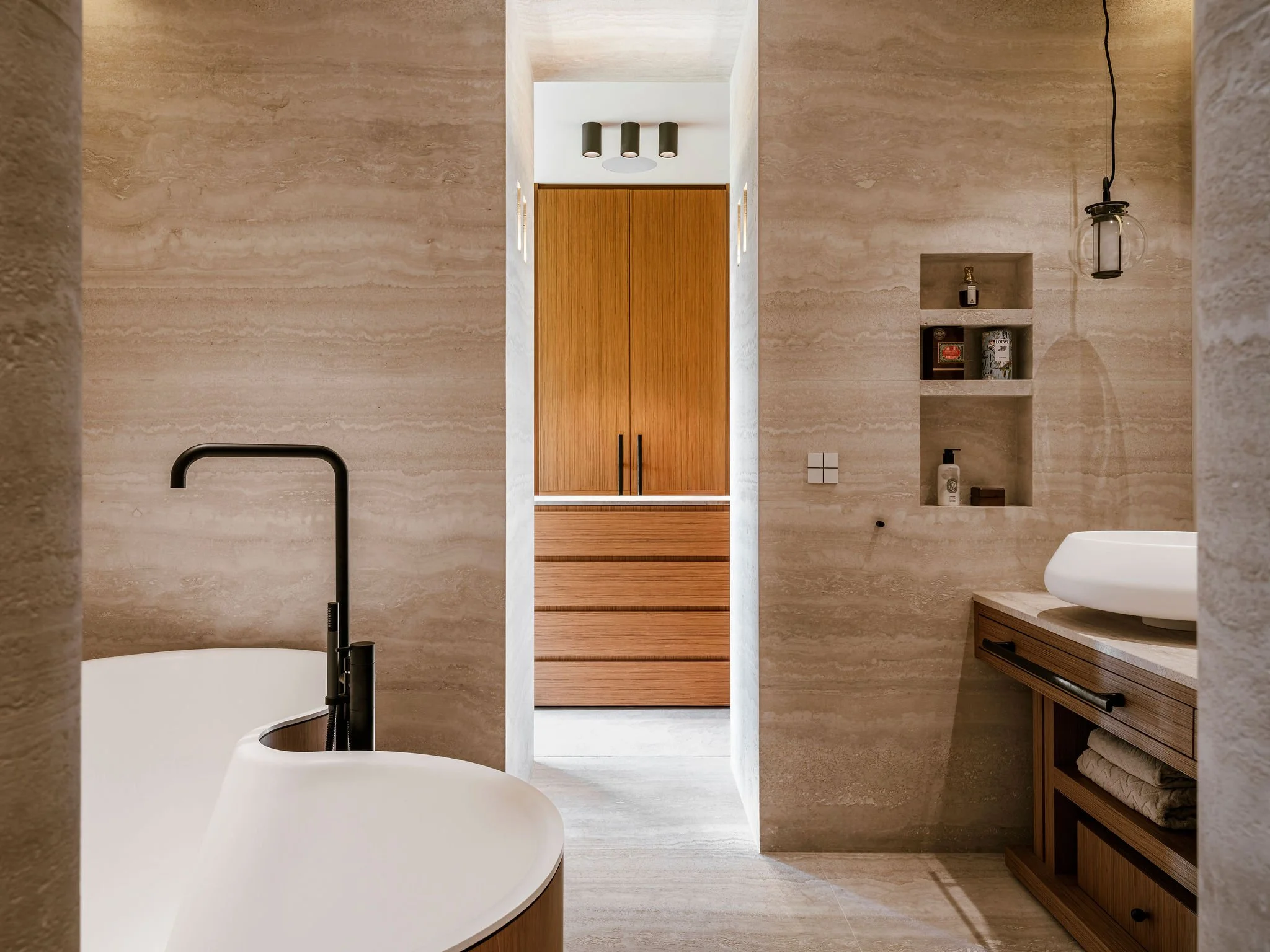 ultra luxury bathroom with beige stone travertine walls, a black Vola faucet, a white Agape bathtub, teak wooden custom cabinetry, and a travertine shelf with high quality toiletries.