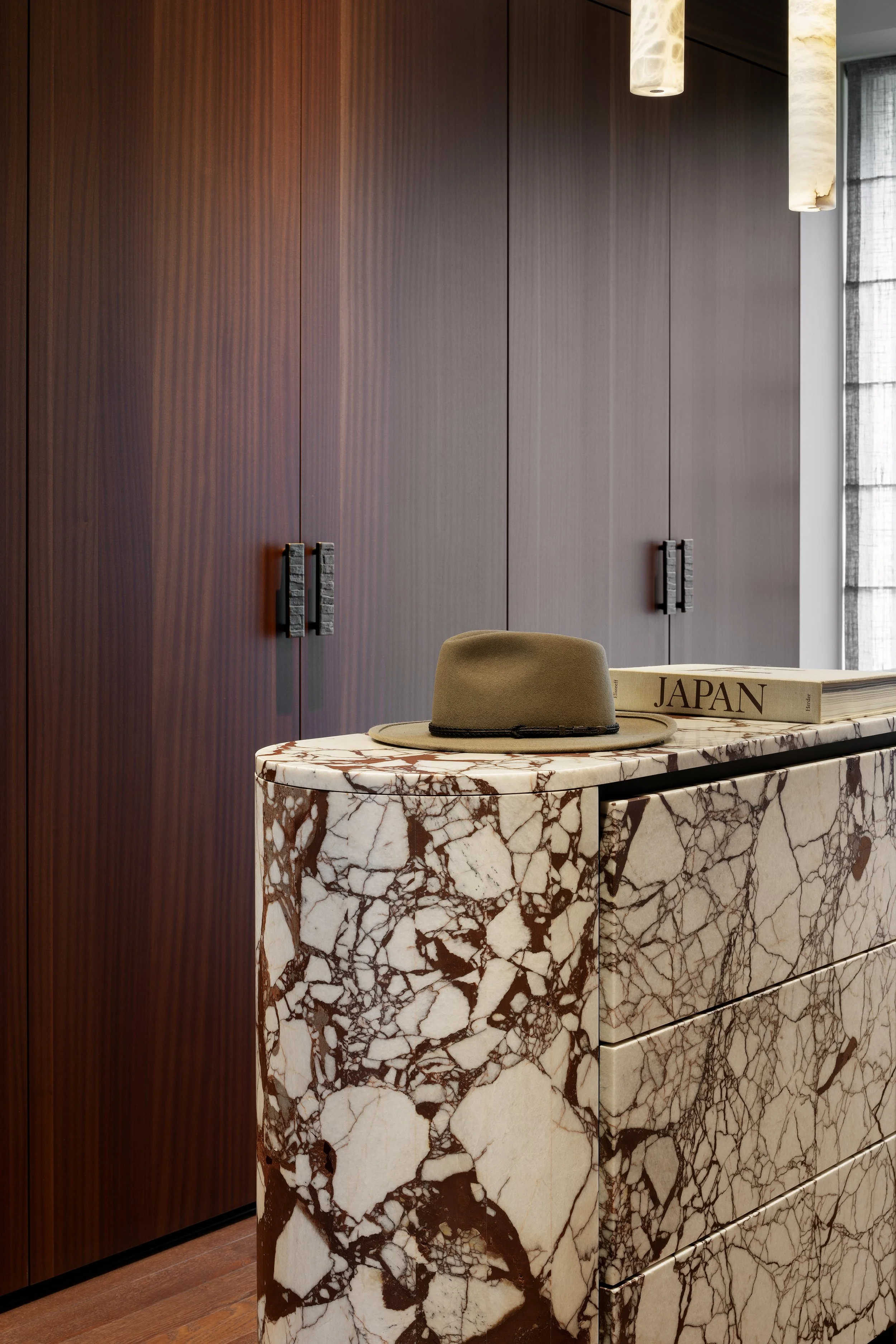 A marble sideboard with reddish brown and white veining, a khaki fedora hat, and a book titled 'JAPAN' on top, against a wooden wardrobe and a window with gray curtains.