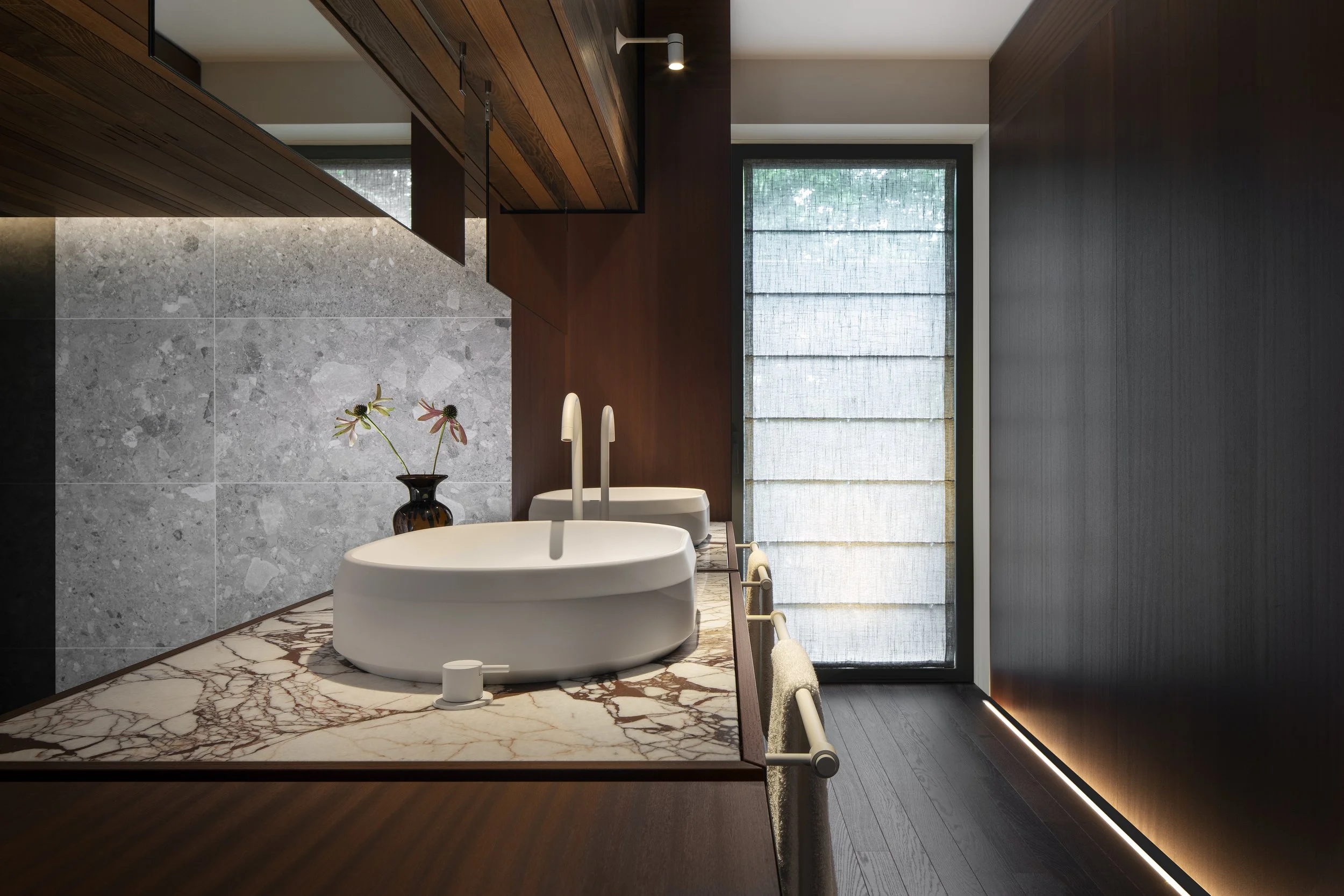 Modern bathroom with dual white vessel sinks from Agape on a marble countertop, a small vase with flowers, and a large window with sheer curtains, dark wood finishes, and minimal decor.