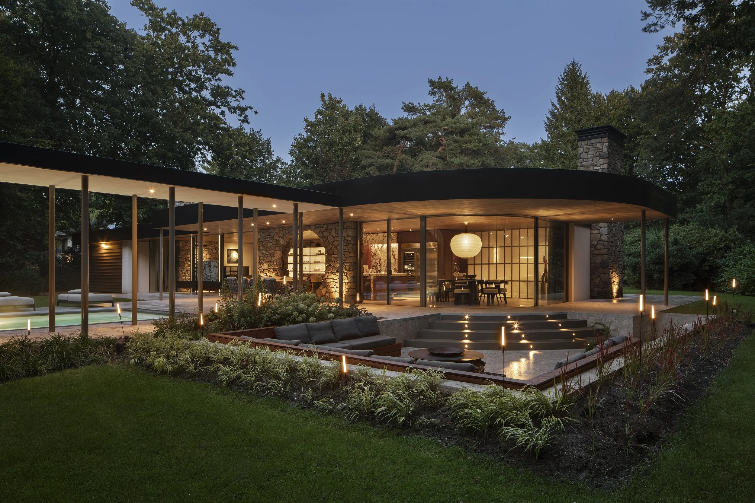 Modern midcentury bungalow with large curved glass windows and outdoor lighting, surrounded by green trees at dusk.