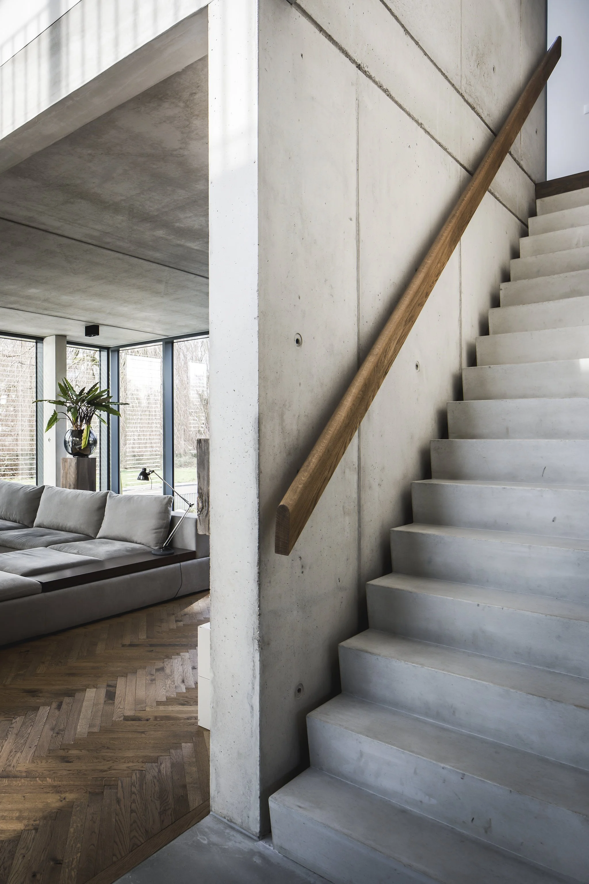Interior of a Loft styled house showing a staircase with a wooden handrail, concrete steps, a concrete wall, and a living room with a gray sofa, wooden flooring, and large windows with a view of the outdoor yard.