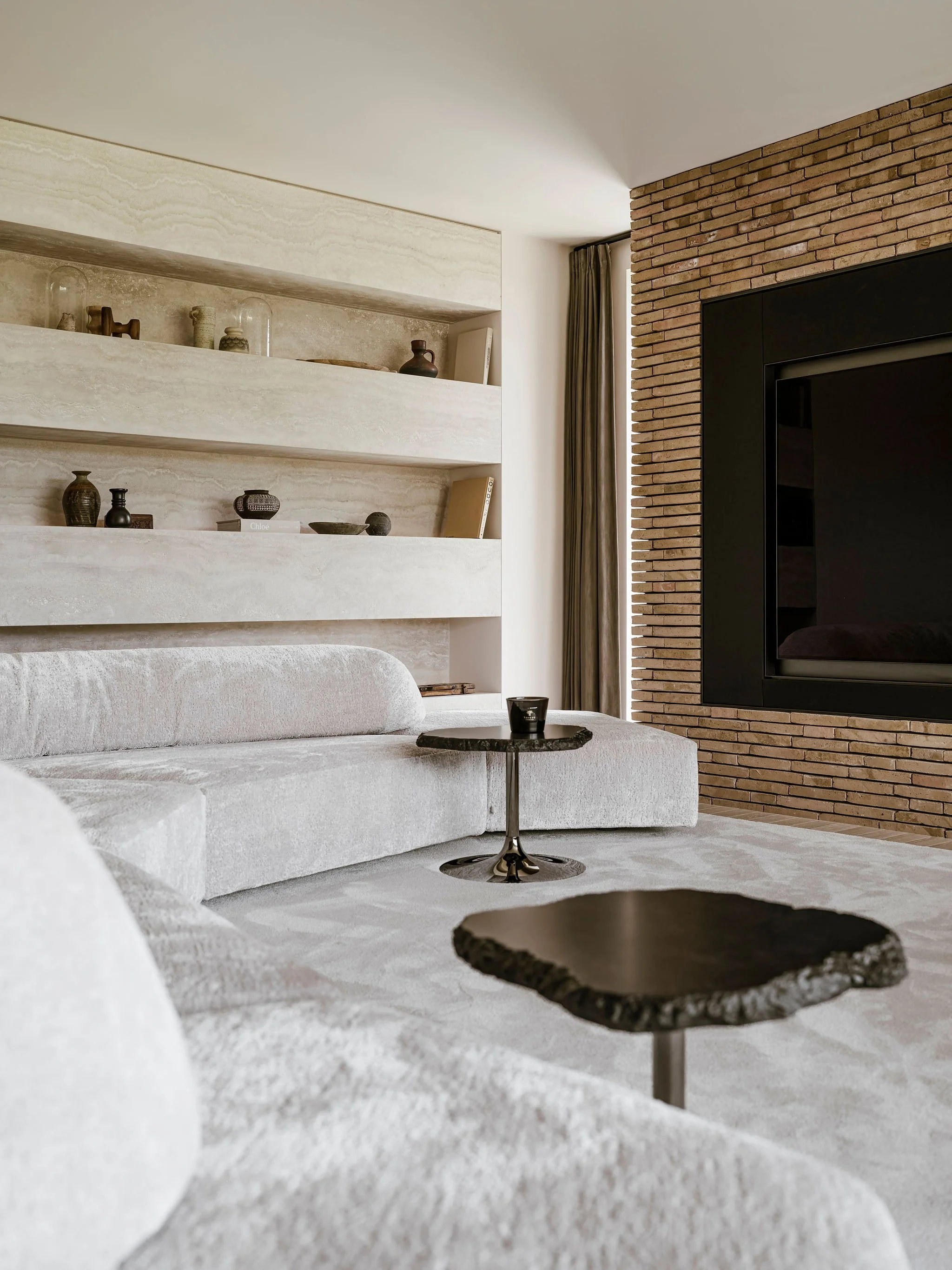 Quiet luxury living room with beige plush Edra sofa, black coffee and side tables, brick accent wall, built-in shelves with decorative objects, and a large TV.