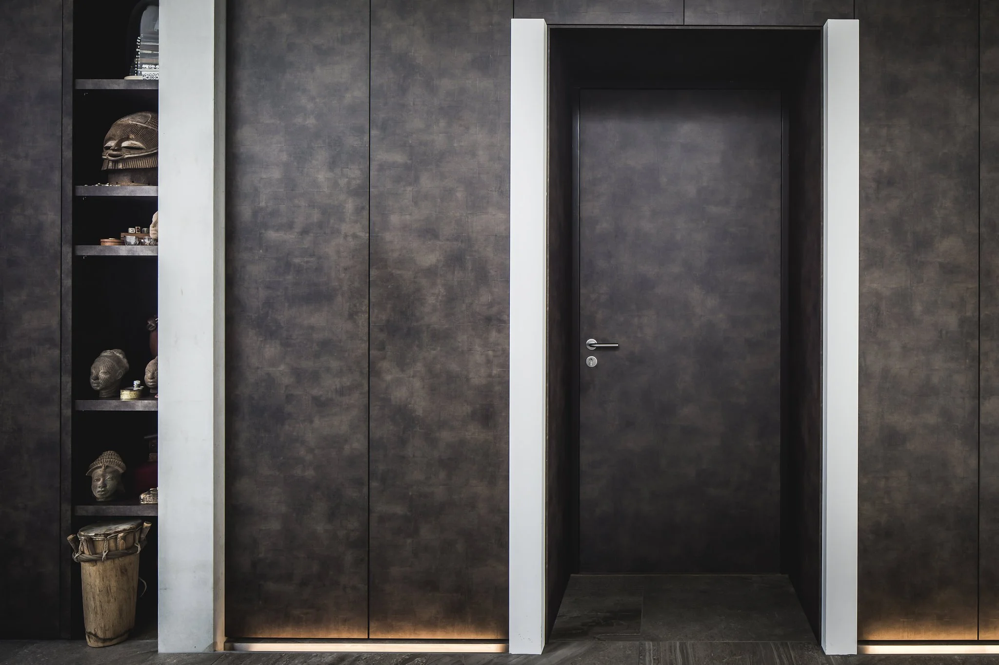 Doorway framed by dark wall leather panels with a small display shelf on the left holding various African artefacts. The floor is dark, and there is a white trim around the doorway. There is LED lighting coming from the ground up.