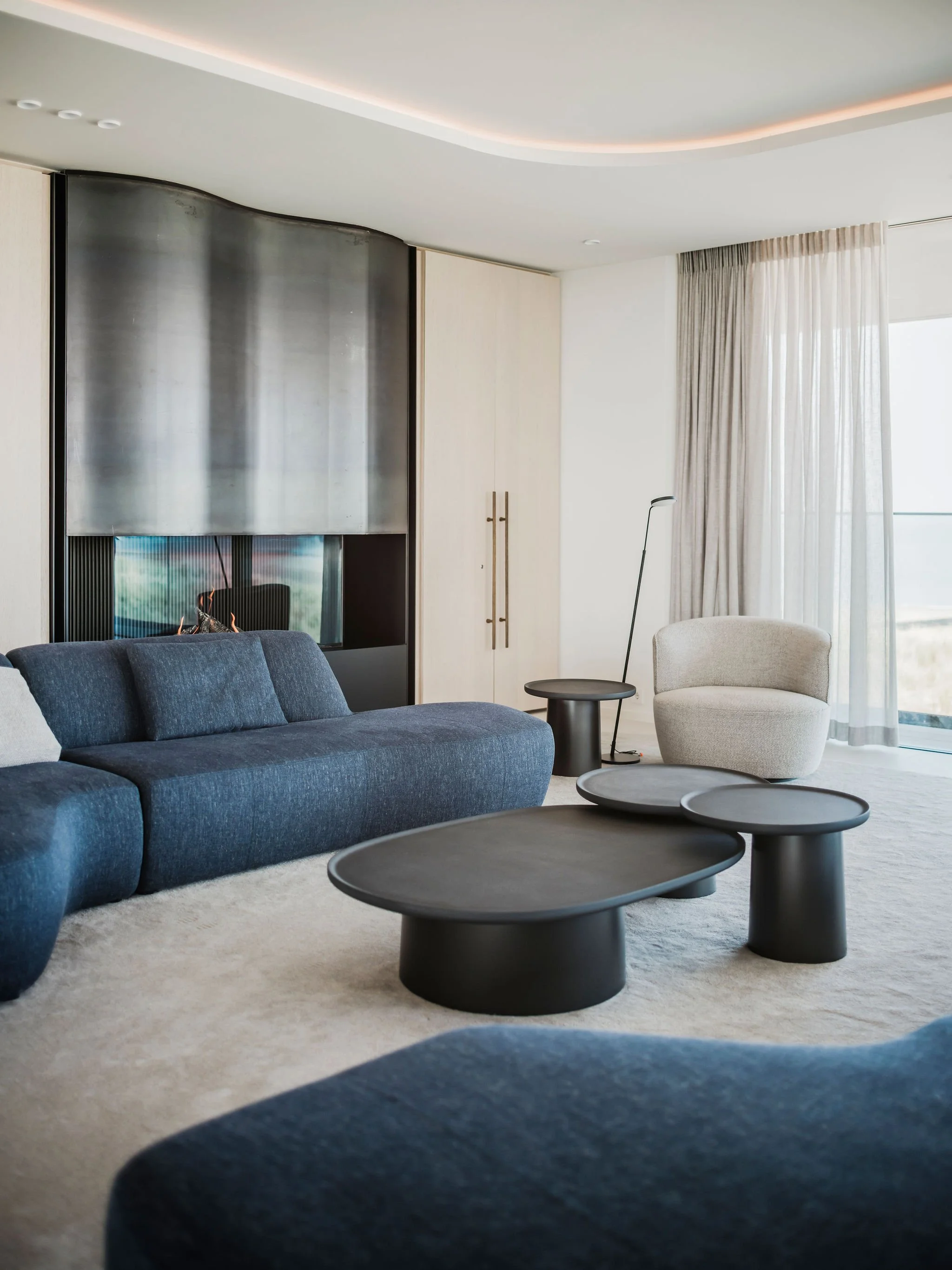 Bespoke  living room with blue sofa, cream armchair, black coffee tables, and a white curtain near large windows and a custom steel fireplace.
