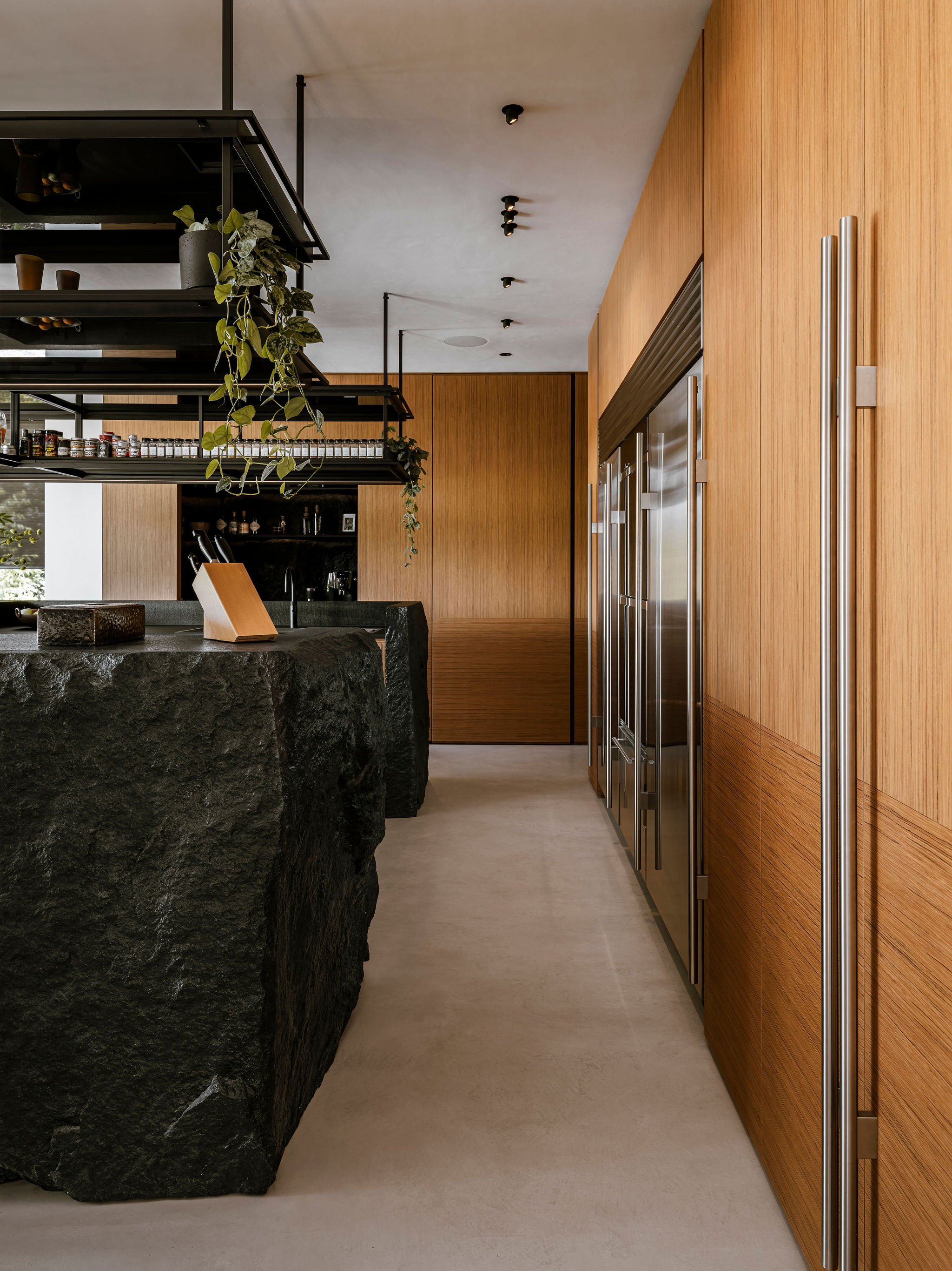 Modern kitchen, custom designed by Guy de Vos with black stone rock island, teak wooden wall panels, stainless steel Subzero refrigerator, and built-in cabinets, with hanging plants and minimal lighting.
