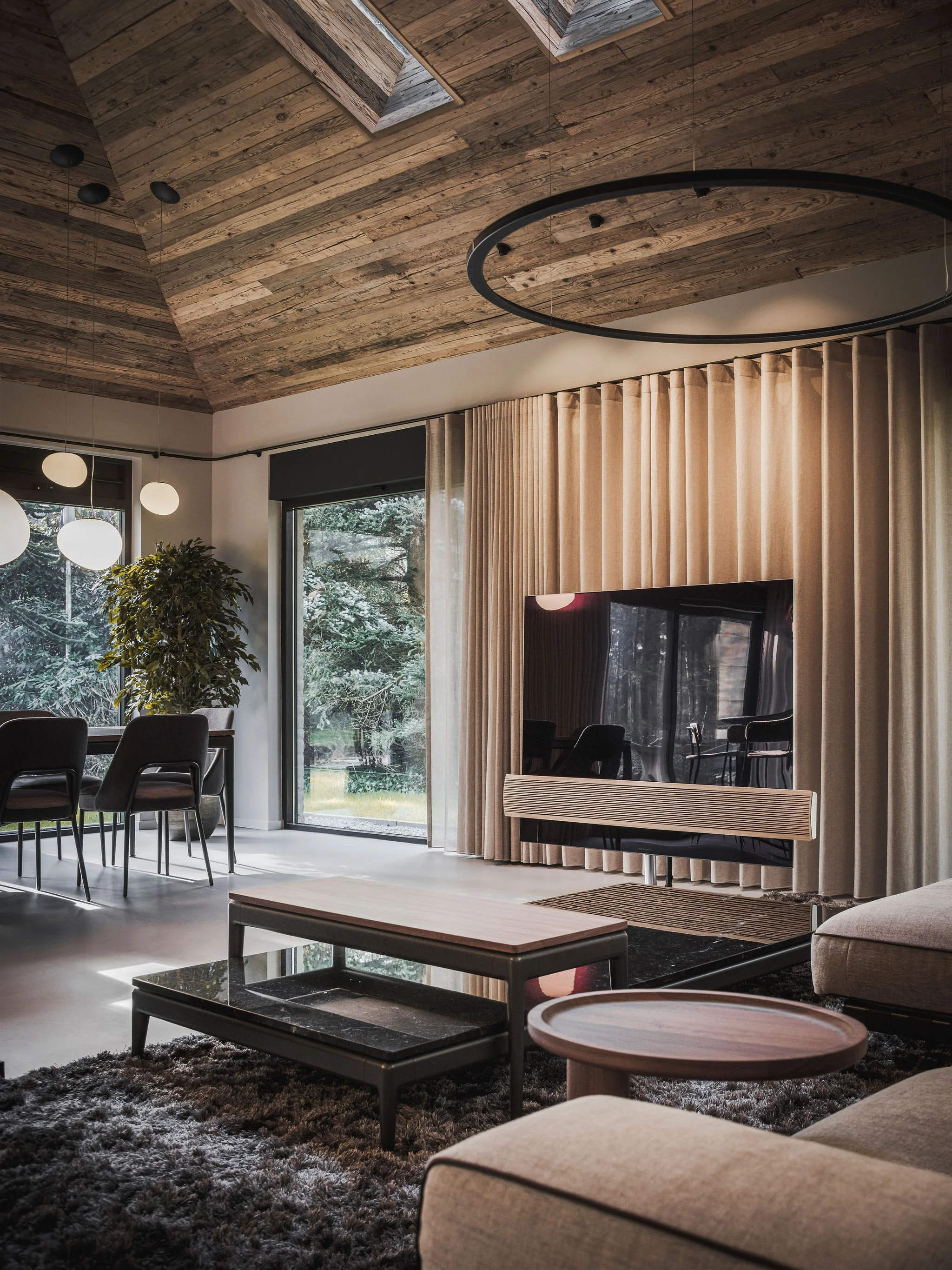 Warm chalet style  living room with wooden ceiling, large windows, grey Flexform sofa, beige curtains, Bang & Olufsen  TV, Flexform coffee tables, and a black modern  chandelier.