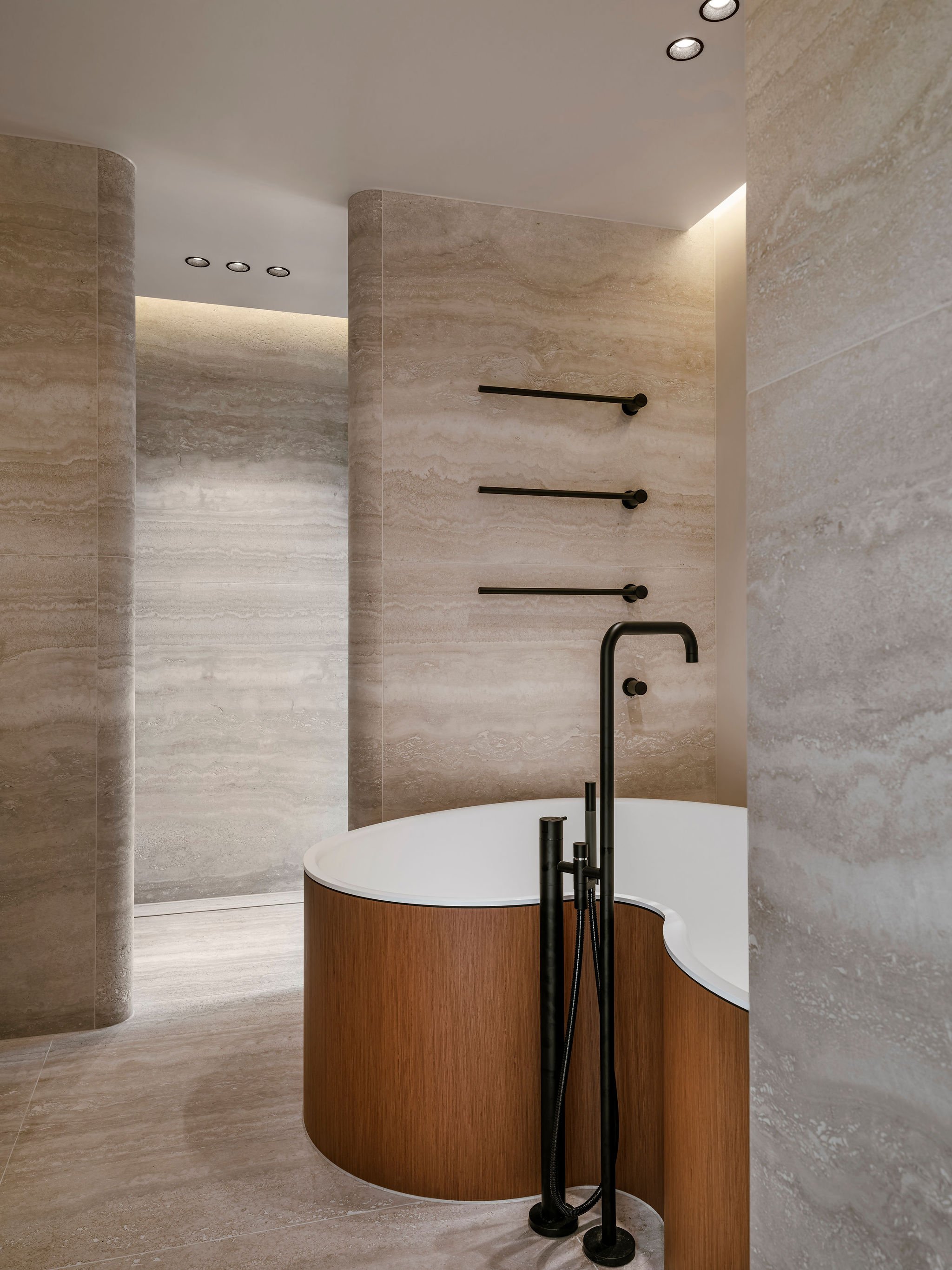 Bespoke quiet luxury bathroom with a freestanding Agape bathtub made of teak wood and white interior, black Vola faucet and shower fixtures, beige Travertine walls and floor, and black Vola towel bars.