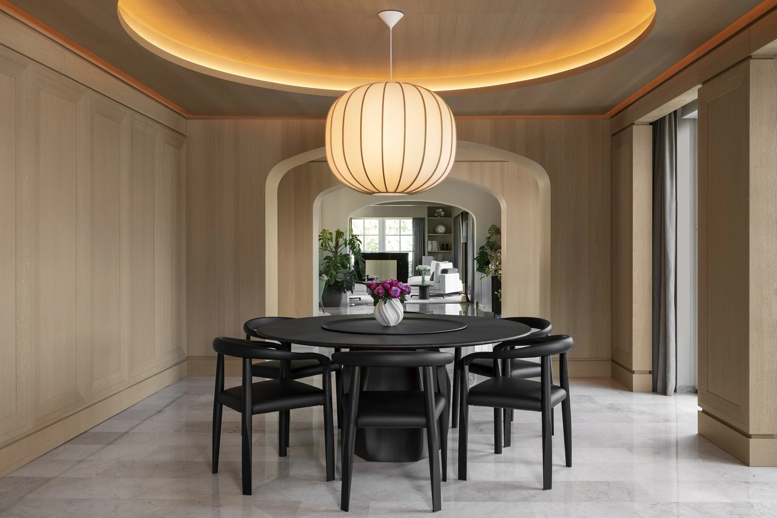 Dining room with a black oval table, six matching black chairs, a white vase with pink flowers, a large hanging paper lantern from Time&Style , Oak wood-paneled walls, and a view into a living room with white furniture and large windows.