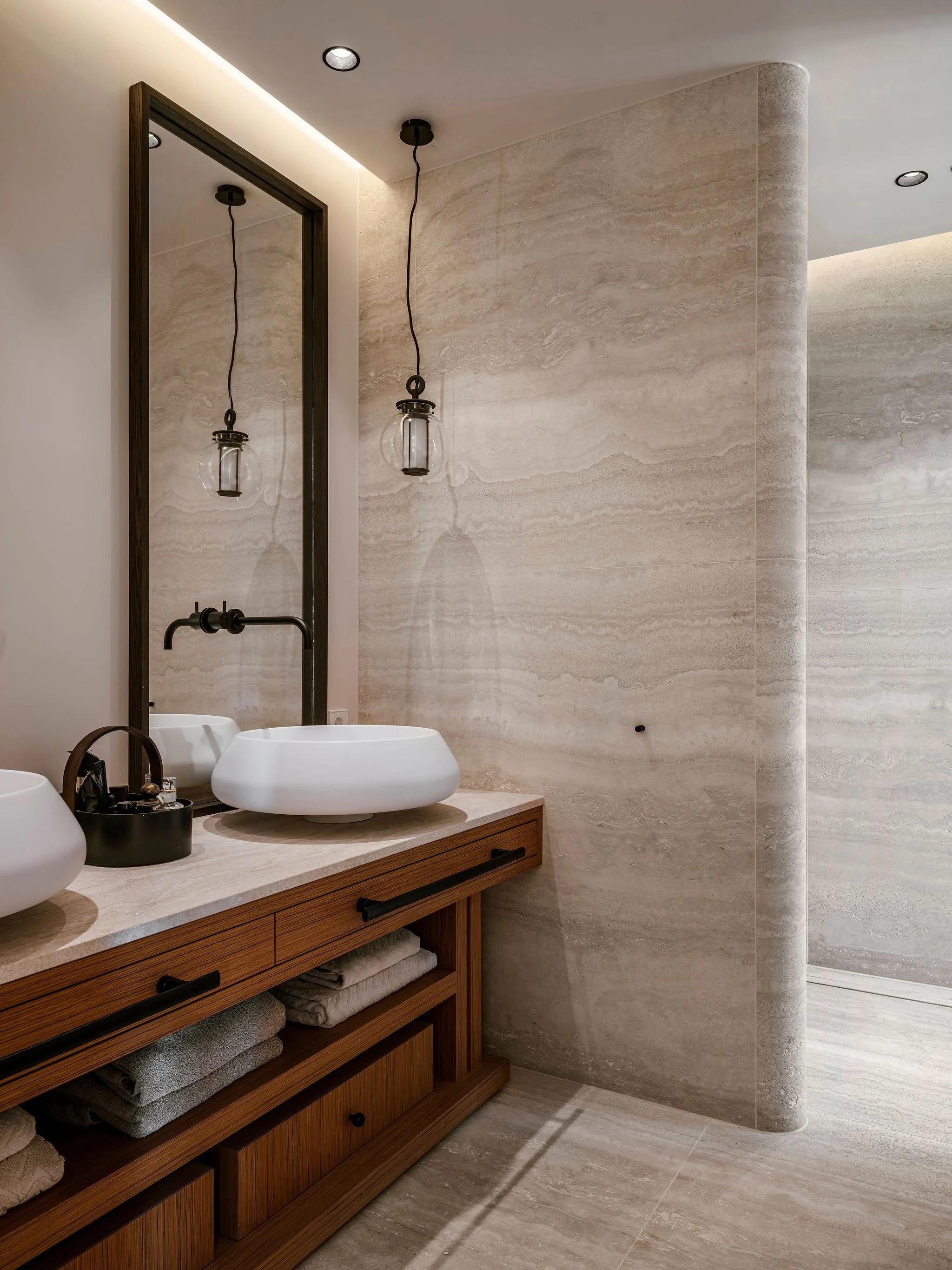 Travertine bathroom with a large mirror, two vessel sinks, a bespoke wooden vanity with open shelves, towels, pendant lights, and a curved natural stone wall.