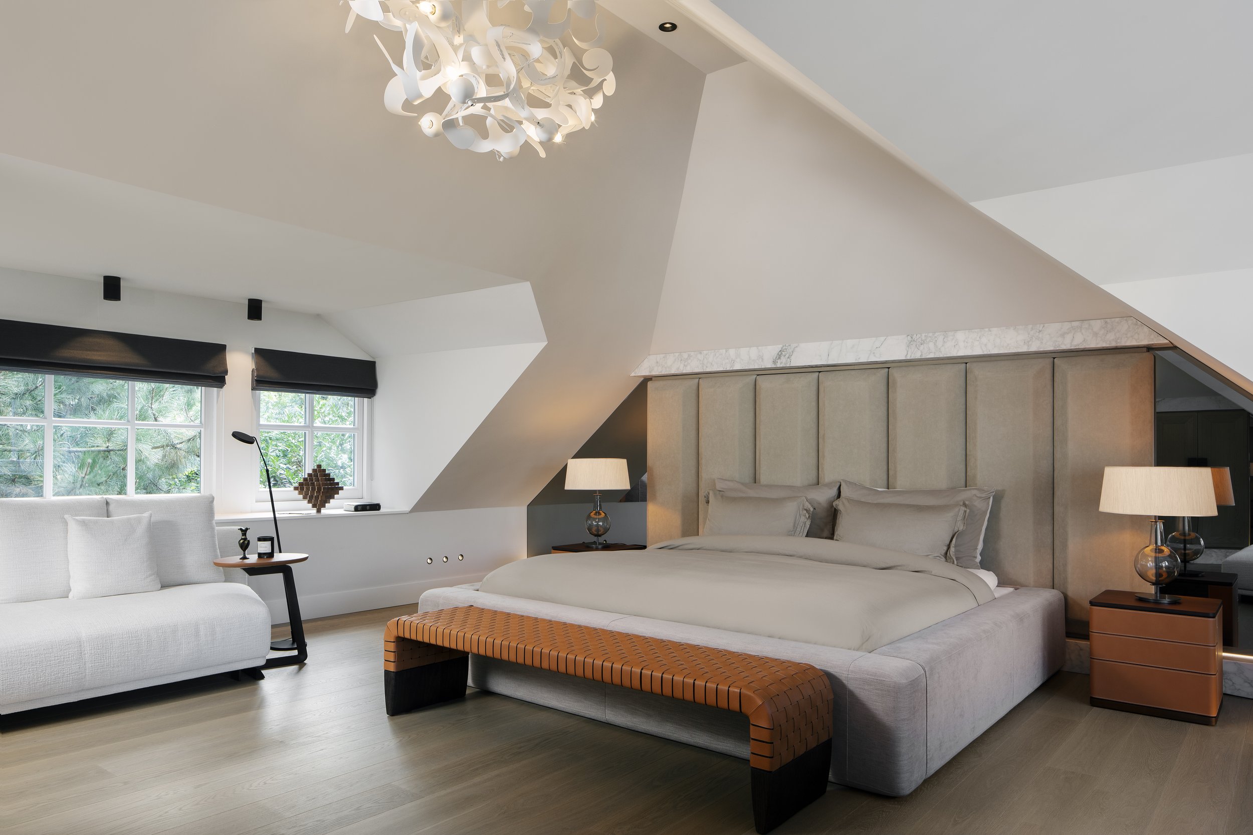 Bedroom with a large bed, beige upholstered headboard, two matching nightstands with lamps, a white sofa, windows with black shades, and unique ceiling design with a chandelier.