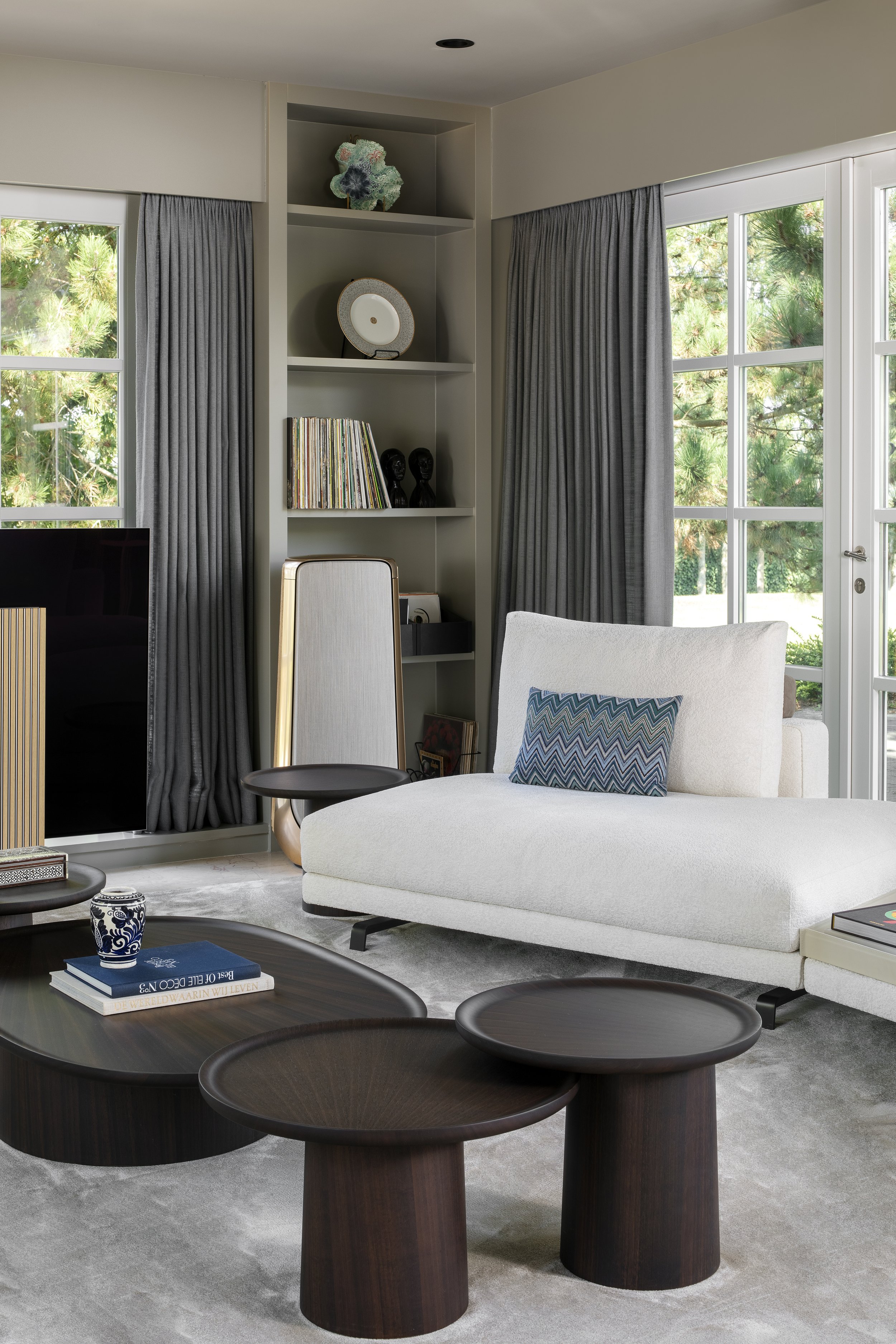 Living room with gray curtains, a white chaise lounge with a zigzag patterned pillow, black round and oval side tables, a B&O TV, and a built-in bookshelf with decorative objects and vinyl records, with large windows revealing green trees outside.
