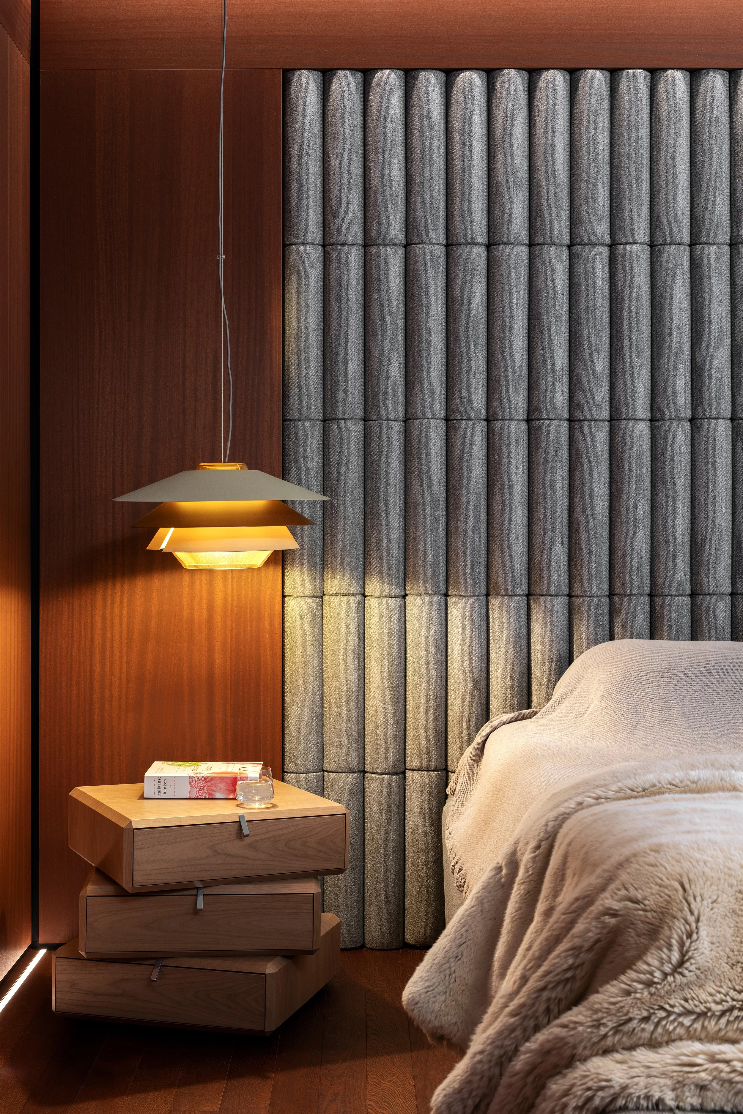 A bedroom corner with a wooden nightstand from Molteni & C holding a book, a glass of water, and a lamp, next to a beige bed with a plush blanket and a padded gray fabric headboard.