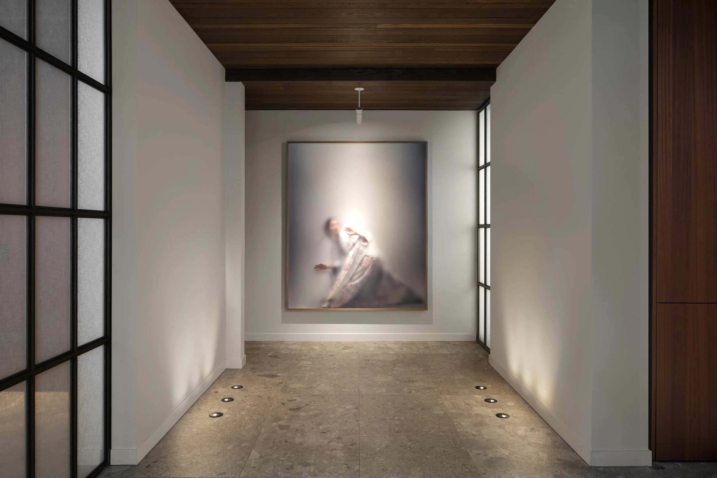 An entrance to a midcentury bungalow  with a artwork of a blurred portrait of a woman in traditional japanese clothing hanging on the wall, illuminated by soft spotlights on the floor, with wooden ceiling and large sliding doors on the sides.