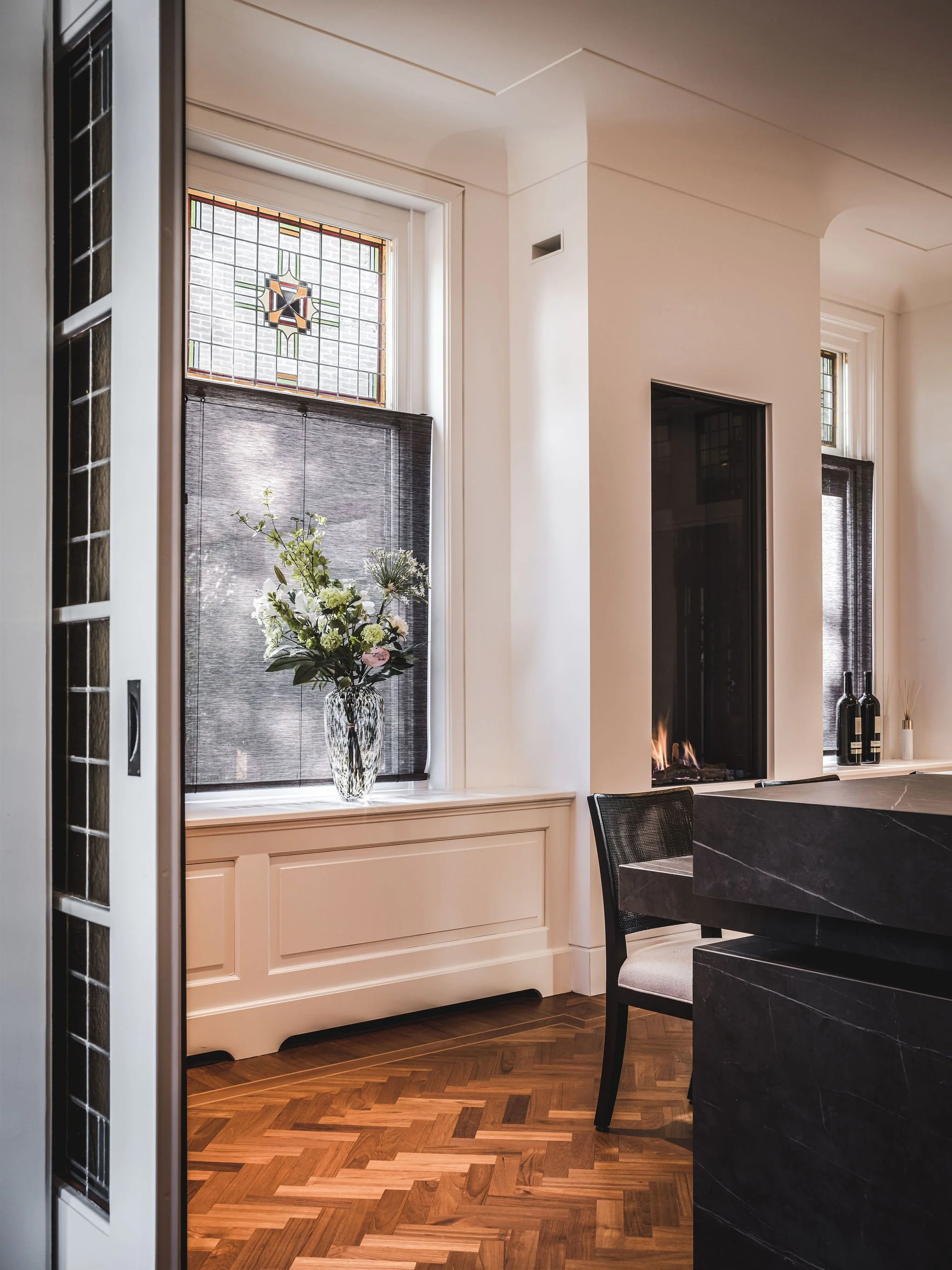 Interior view of a living space with a window featuring stained glass and black window shades, a vase with flowers on the window sill, parquet wood flooring, a black marble kitchen island, and a built-in fireplace.
