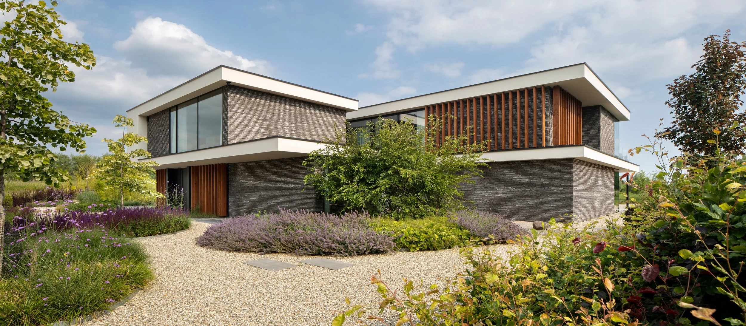 Modern two-story house with large glass windows, gray brick exterior, and wooden accents, surrounded by landscaped garden with purple and green plants, on a sunny day with partly cloudy sky.
