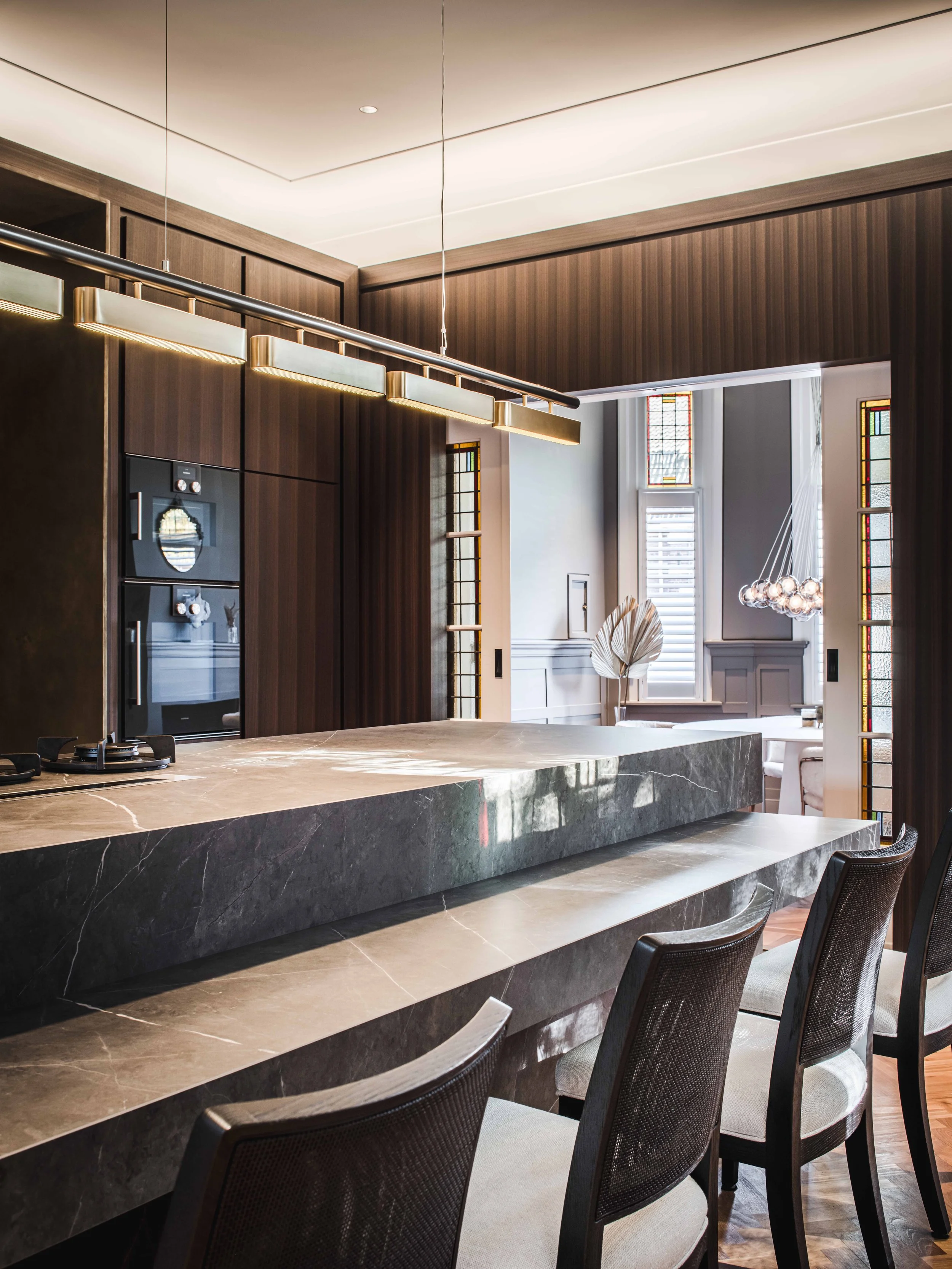 Warm bespoke kitchen with marble island, Maxalto bar chairs, wooden dark  cabinetry, and decorative lighting fixtures.
