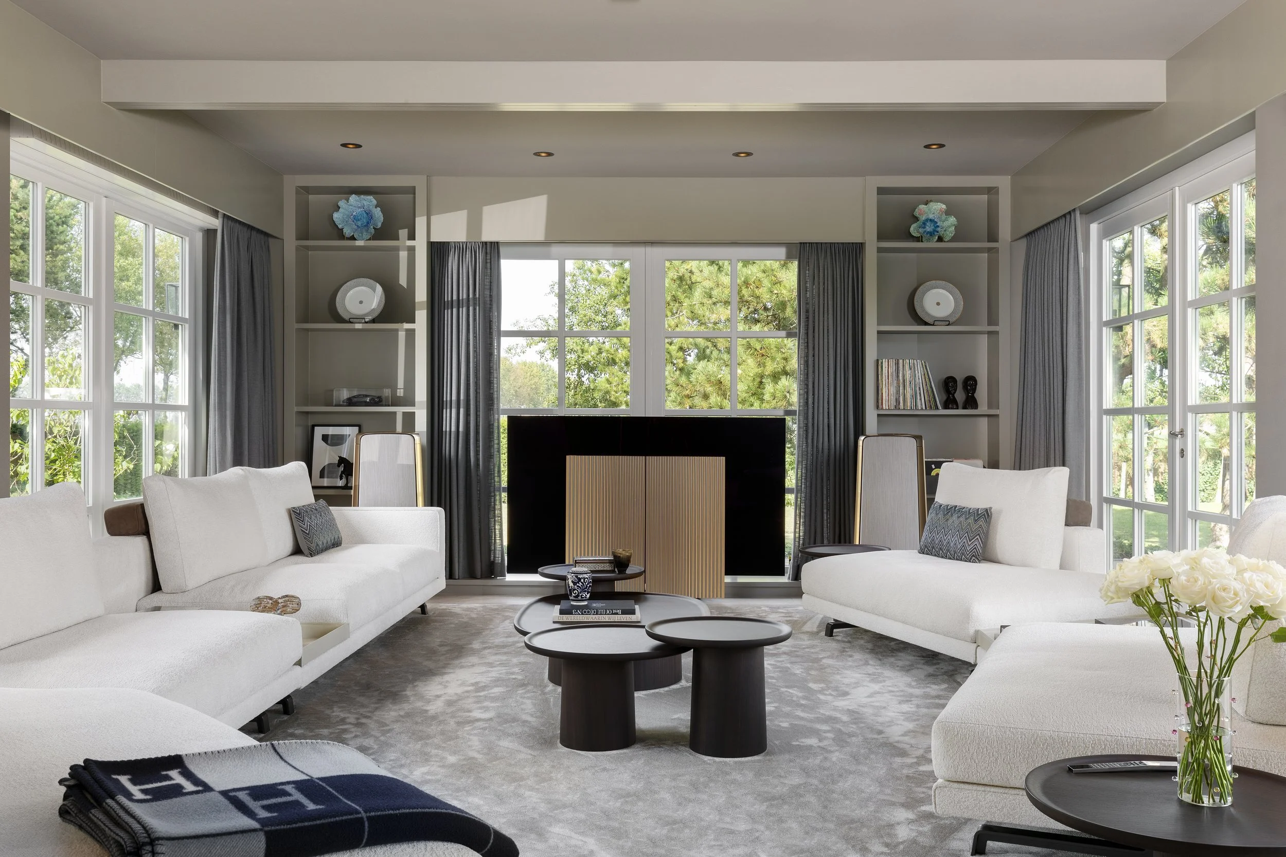 Living room with large windows, white sofas, black coffee tables, Bang & Olufsen Tv & Speakers, and built-in shelves with decorative items and books.